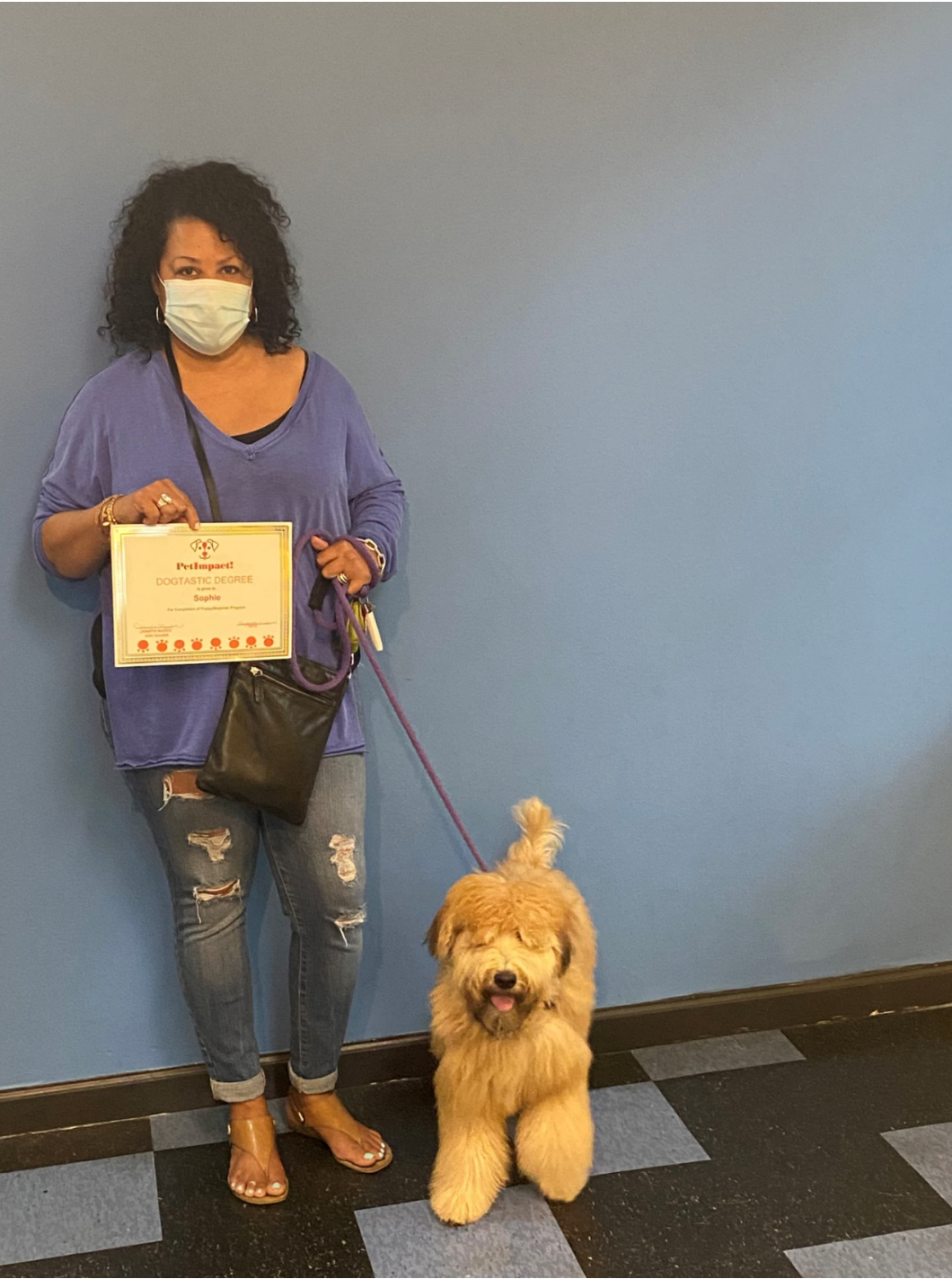 Woman in mask holds a certificate, standing with a fluffy beige dog on a leash in front of a blue wall.
