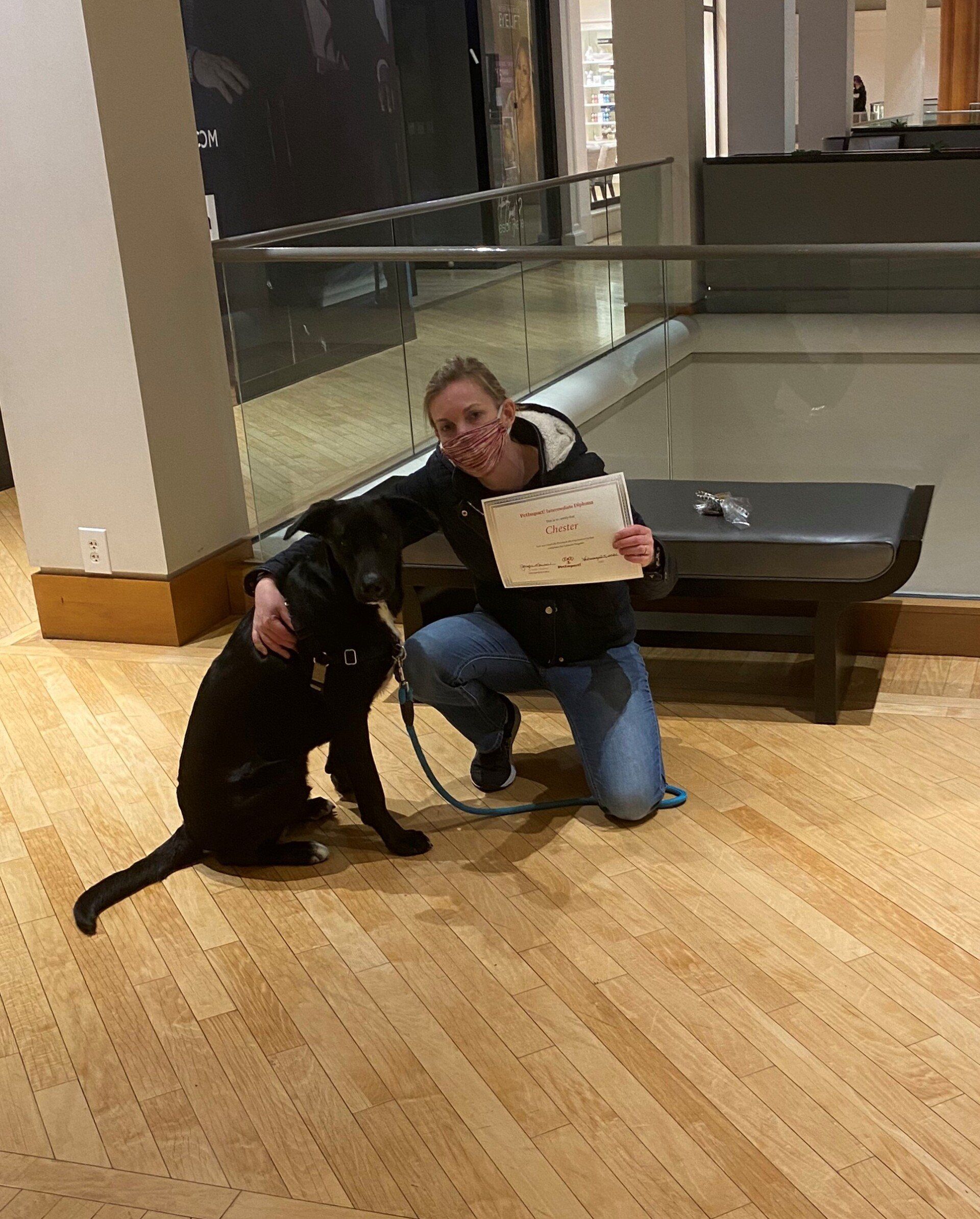 Woman kneeling, hugging black dog, holding paper. Inside building with wooden floor and bench.