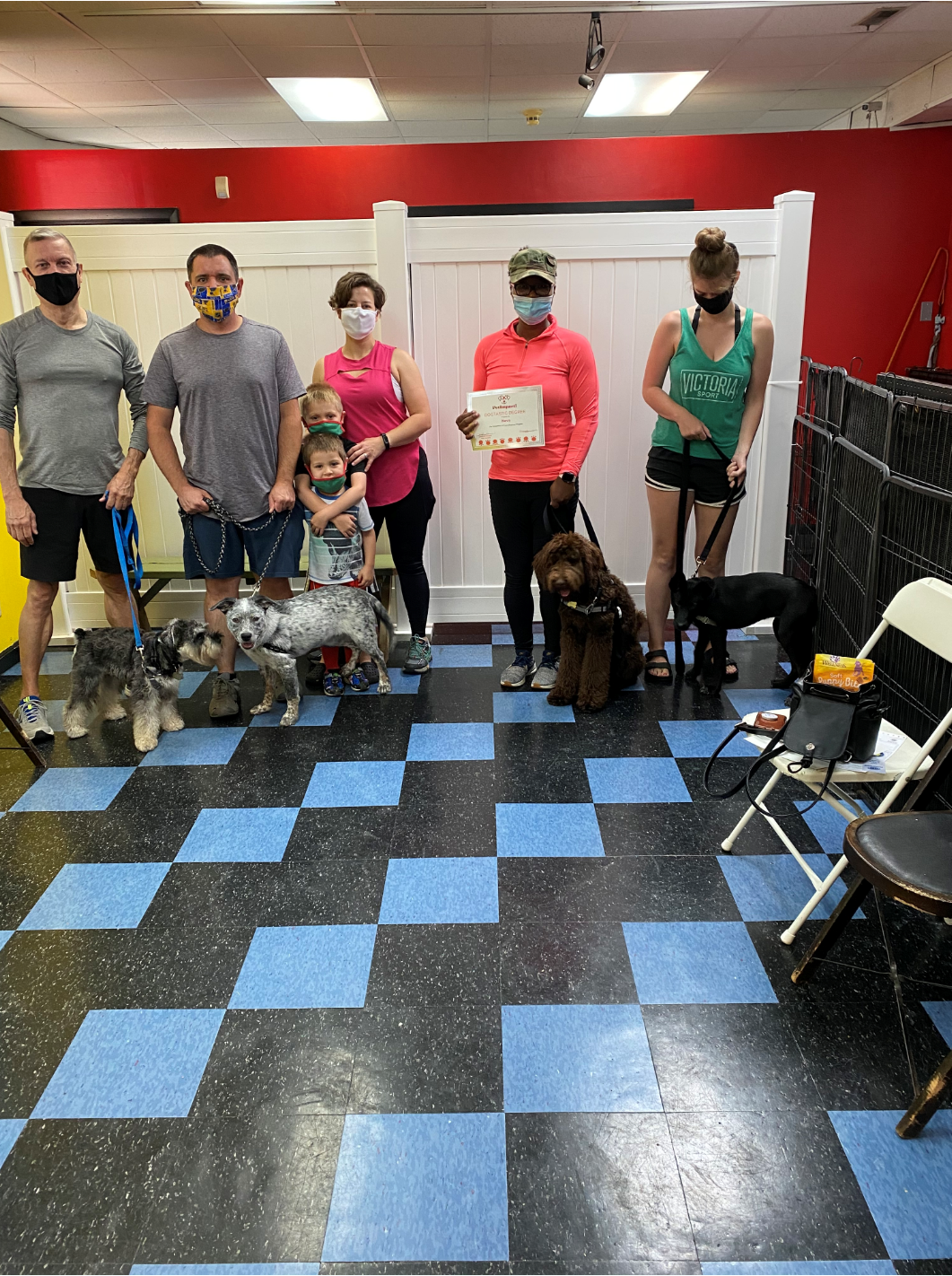 People with dogs in a dog training class, holding certificates. Indoor setting with blue and black checkered floor.