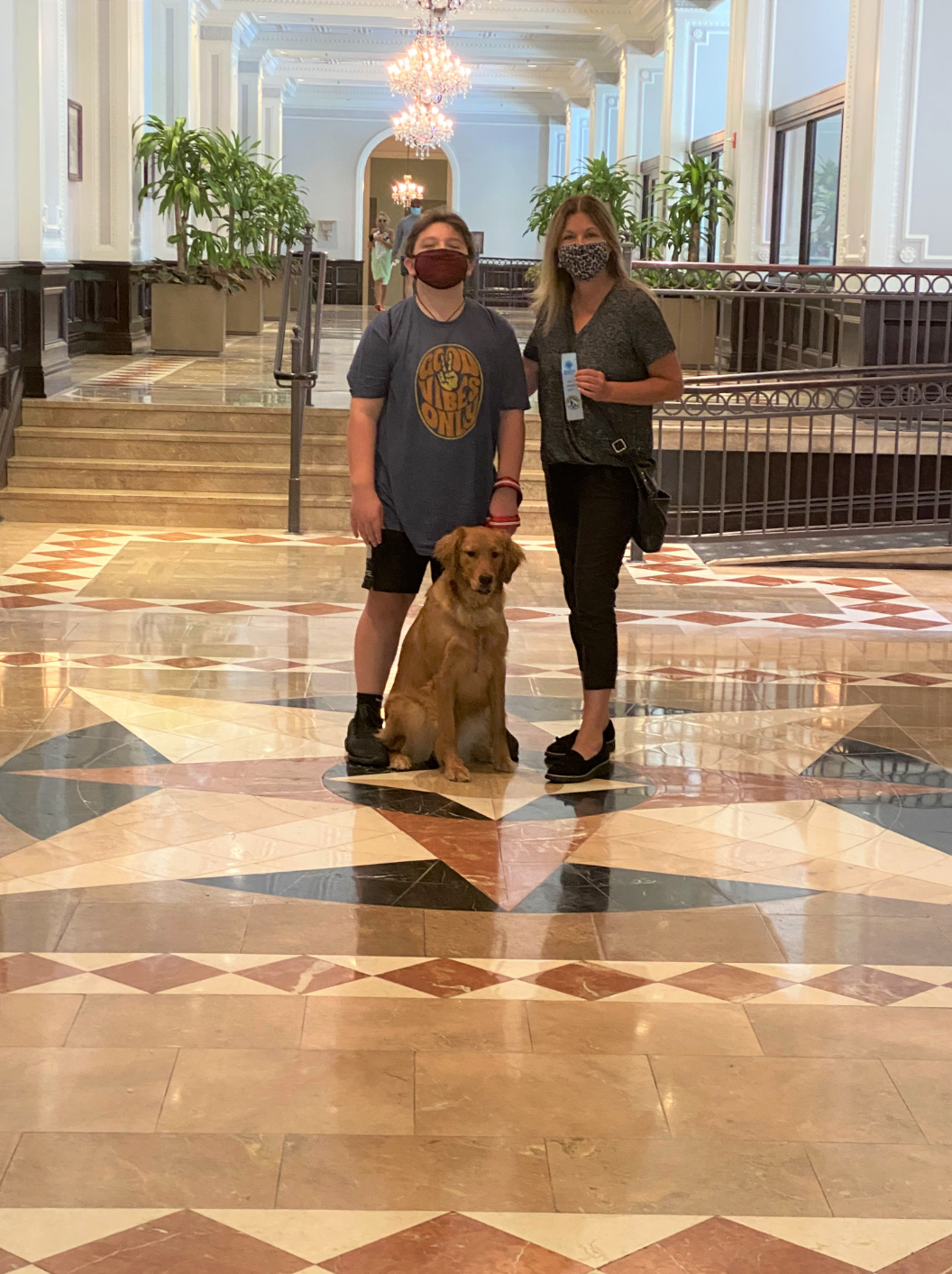 Two people and a golden retriever pose in a lobby. They wear masks. Colorful patterned floor.