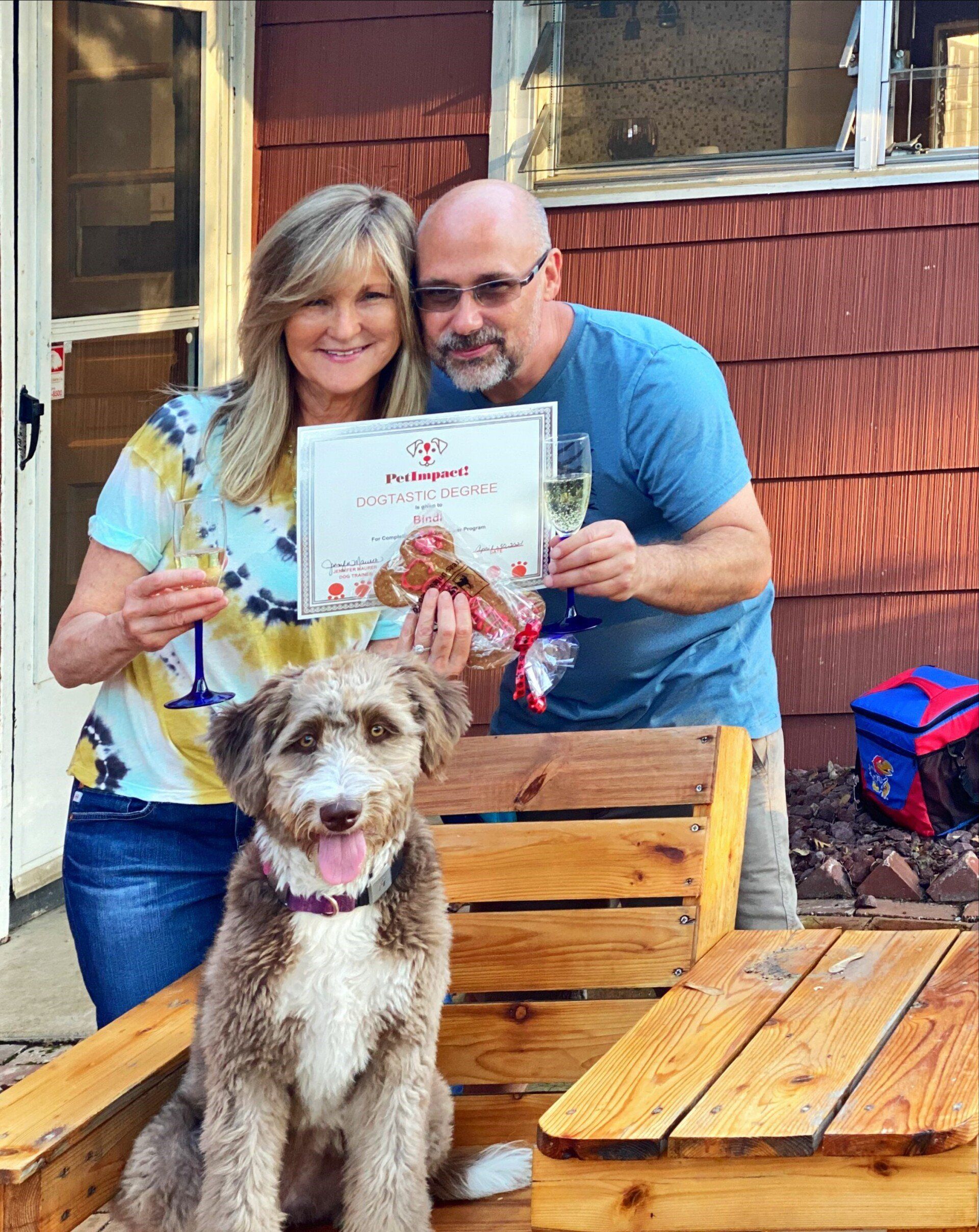 Couple holding certificate and champagne, smiling, with dog in front of wooden crates.