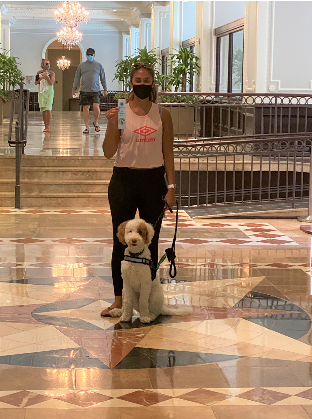 Woman in pink top, mask, and black pants holds a product, standing with a dog on a leash in a building with marble floors.