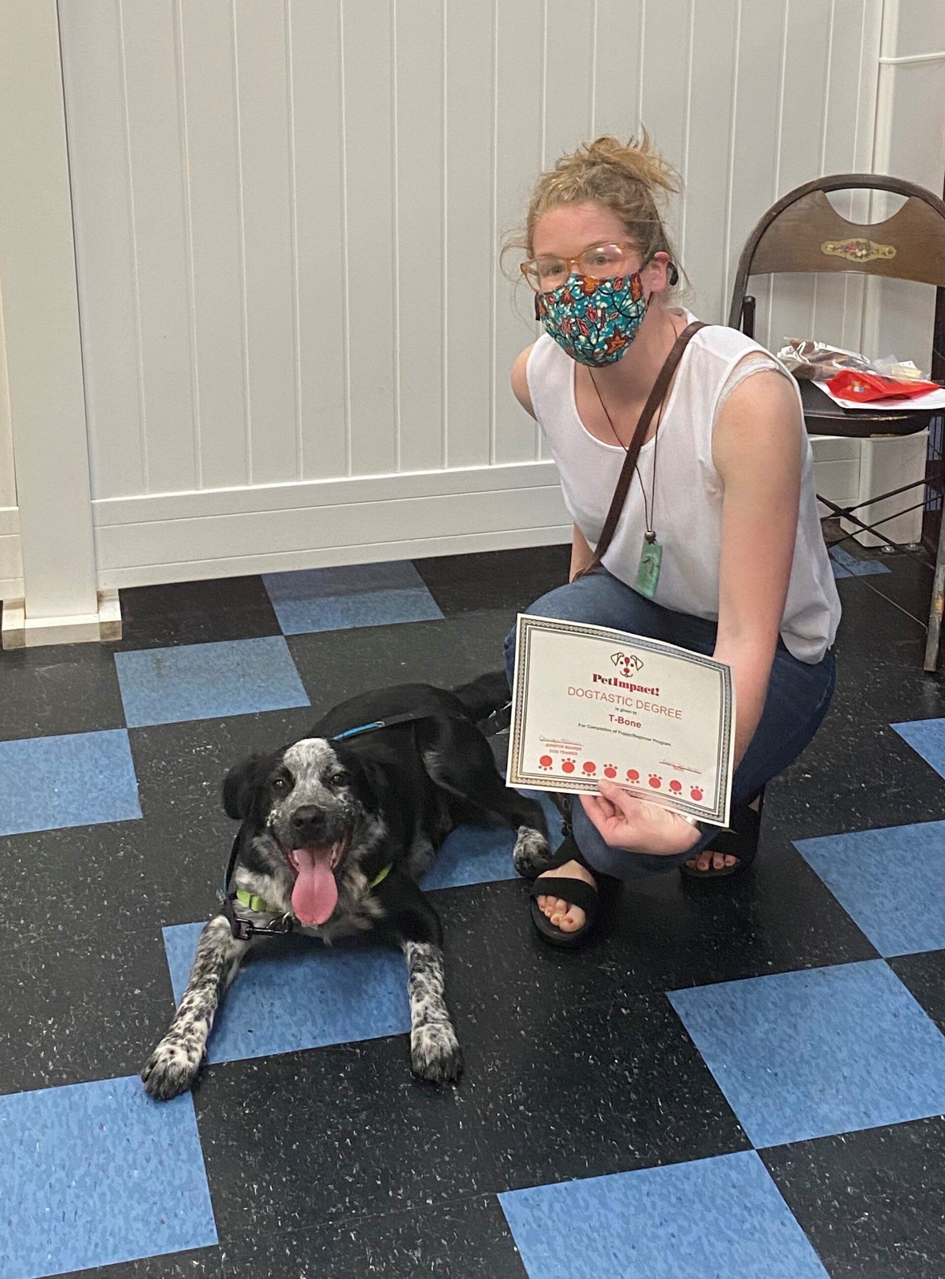 Woman in mask kneels with dog. Woman holds card. Dog has spotted coat, tongue out. Setting is tiled floor.