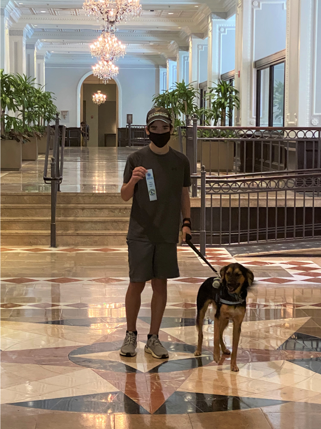 Man in mask and dog in grand hallway; man holds ribbon.