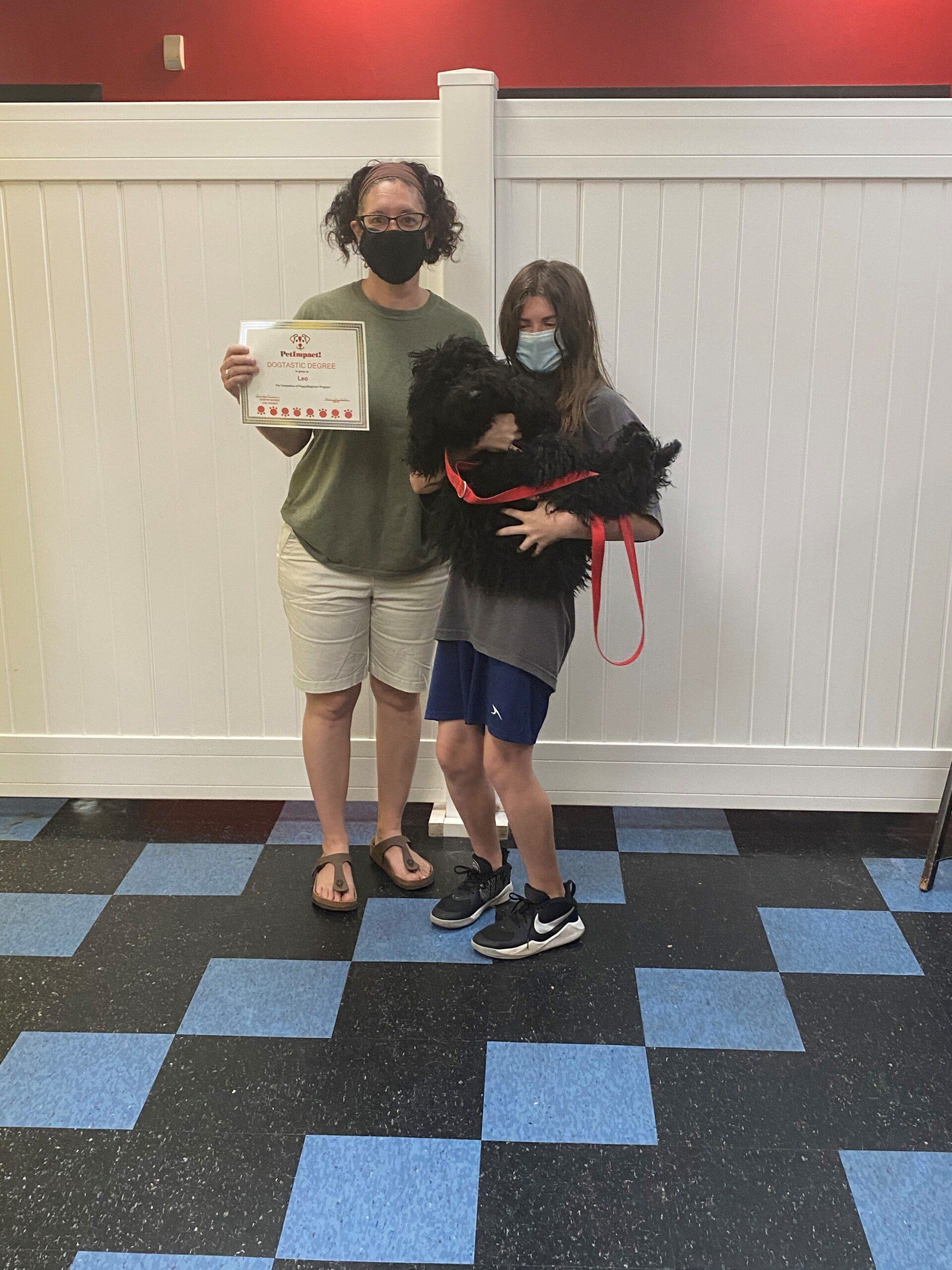 Woman and child in masks with a black dog and certificate, in front of a white wall, on a blue and black floor.