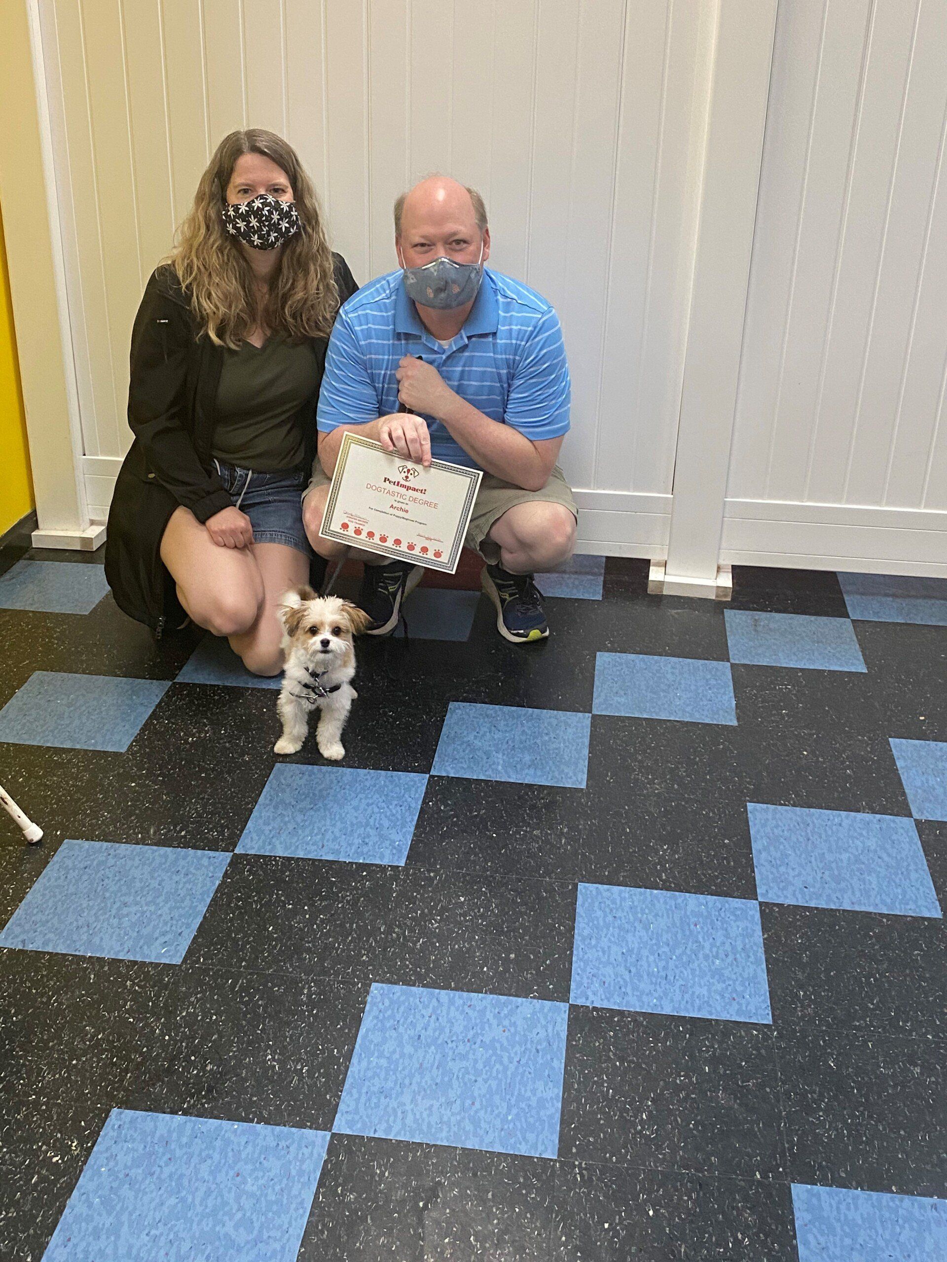 Couple crouching with a small dog and a sign on a checkered floor, all wearing face masks.