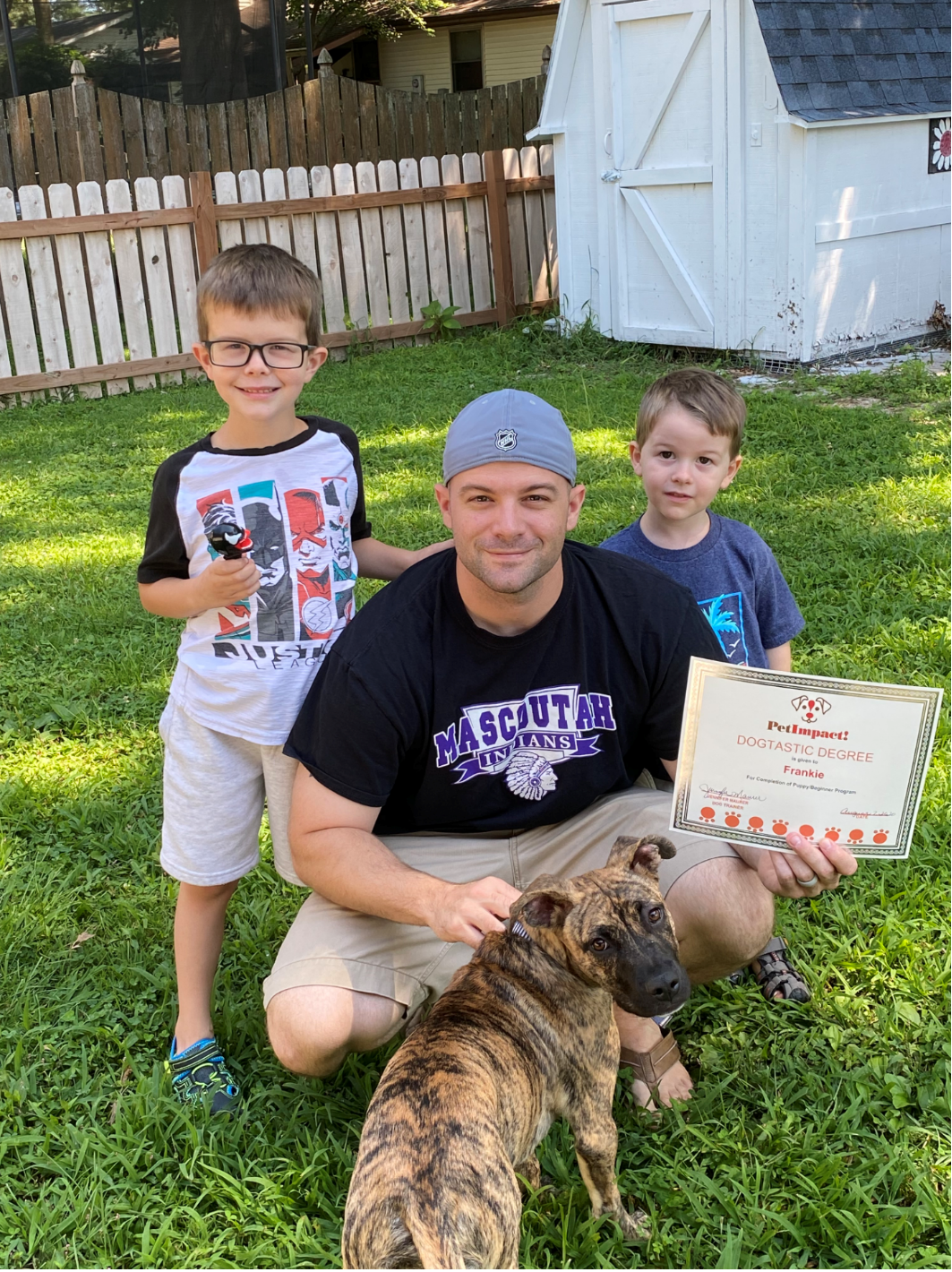 Man and two boys with dog, holding certificate. They are outside near fence and shed.