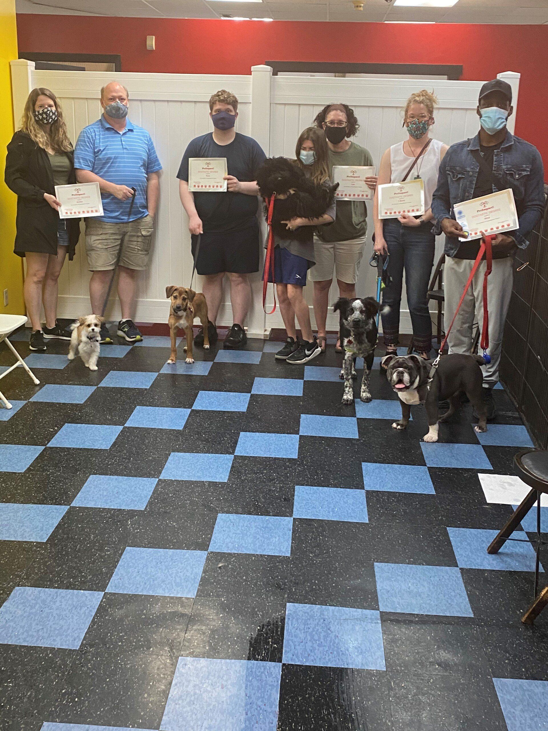 Group of people with dogs holding certificates in a checkered-floor room. People wear masks. Dogs are of various breeds.