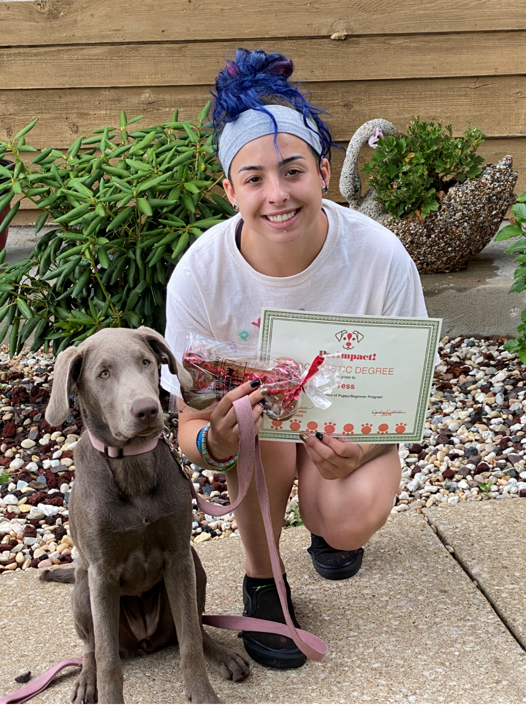 Woman and silver dog kneeling together holding a treat and certificate outside.