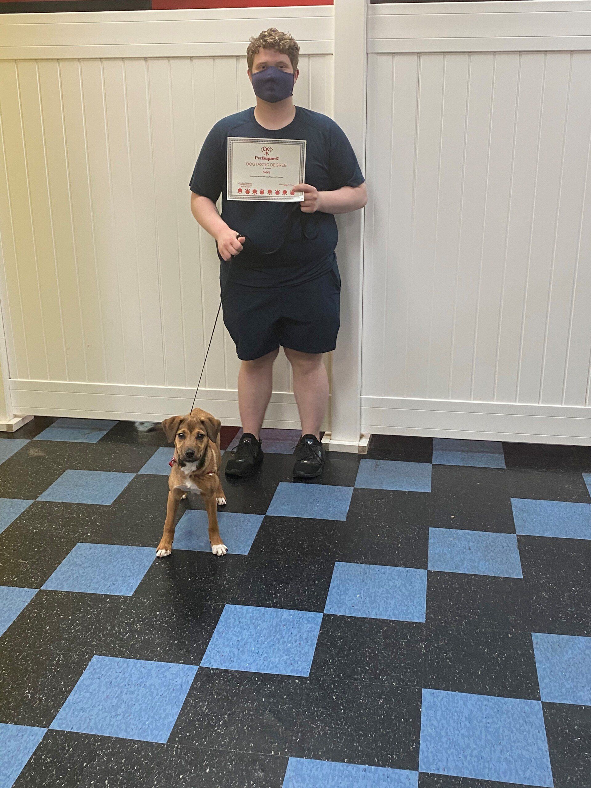 Person in blue shirt and shorts, holding certificate, stands with a puppy on a leash on checkered floor.