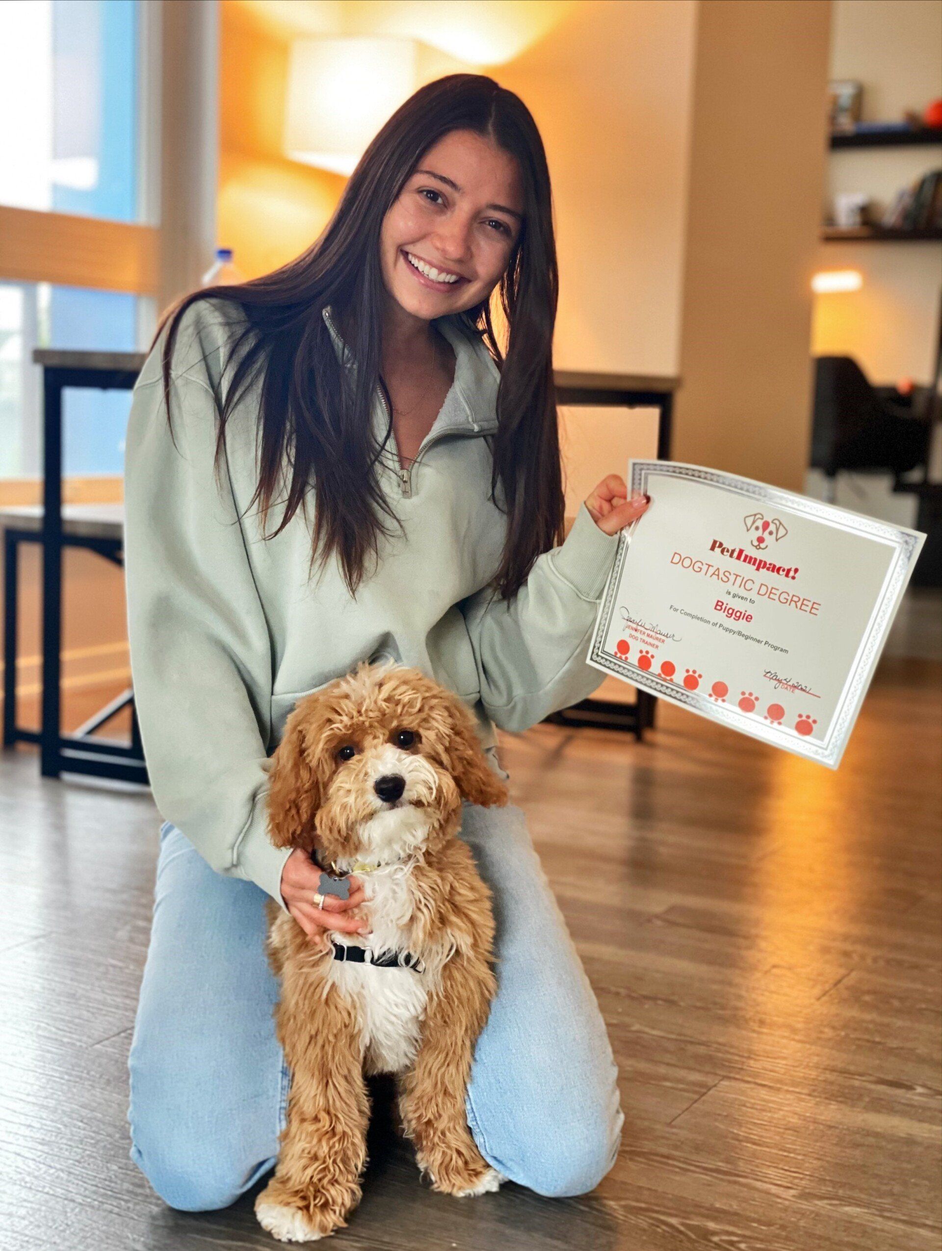 Woman kneeling with a puppy; holding a certificate. She smiles in a light green sweatshirt and jeans. Indoor setting.