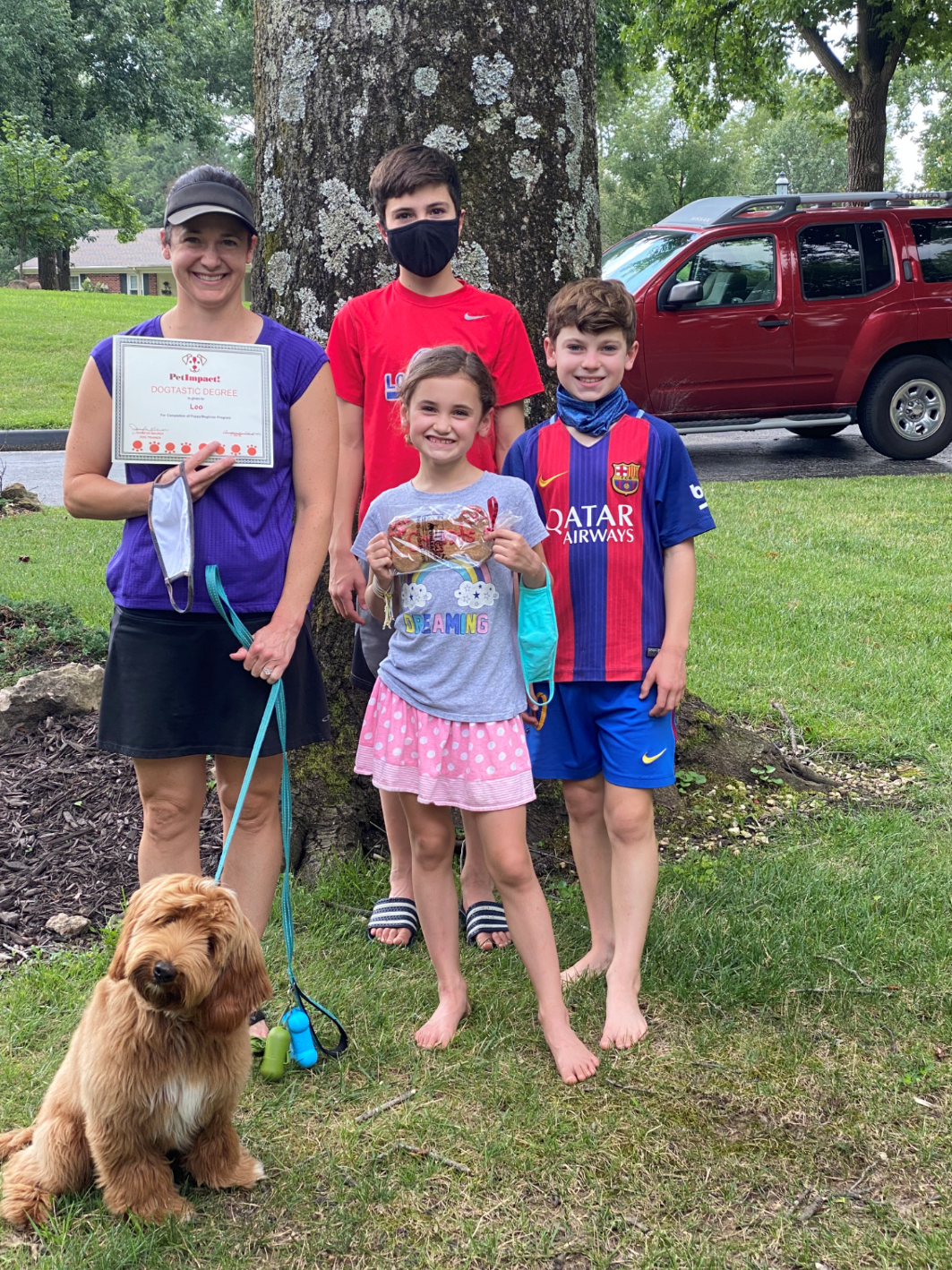 Family with a dog in front of a tree, holding a certificate. Red car in background.