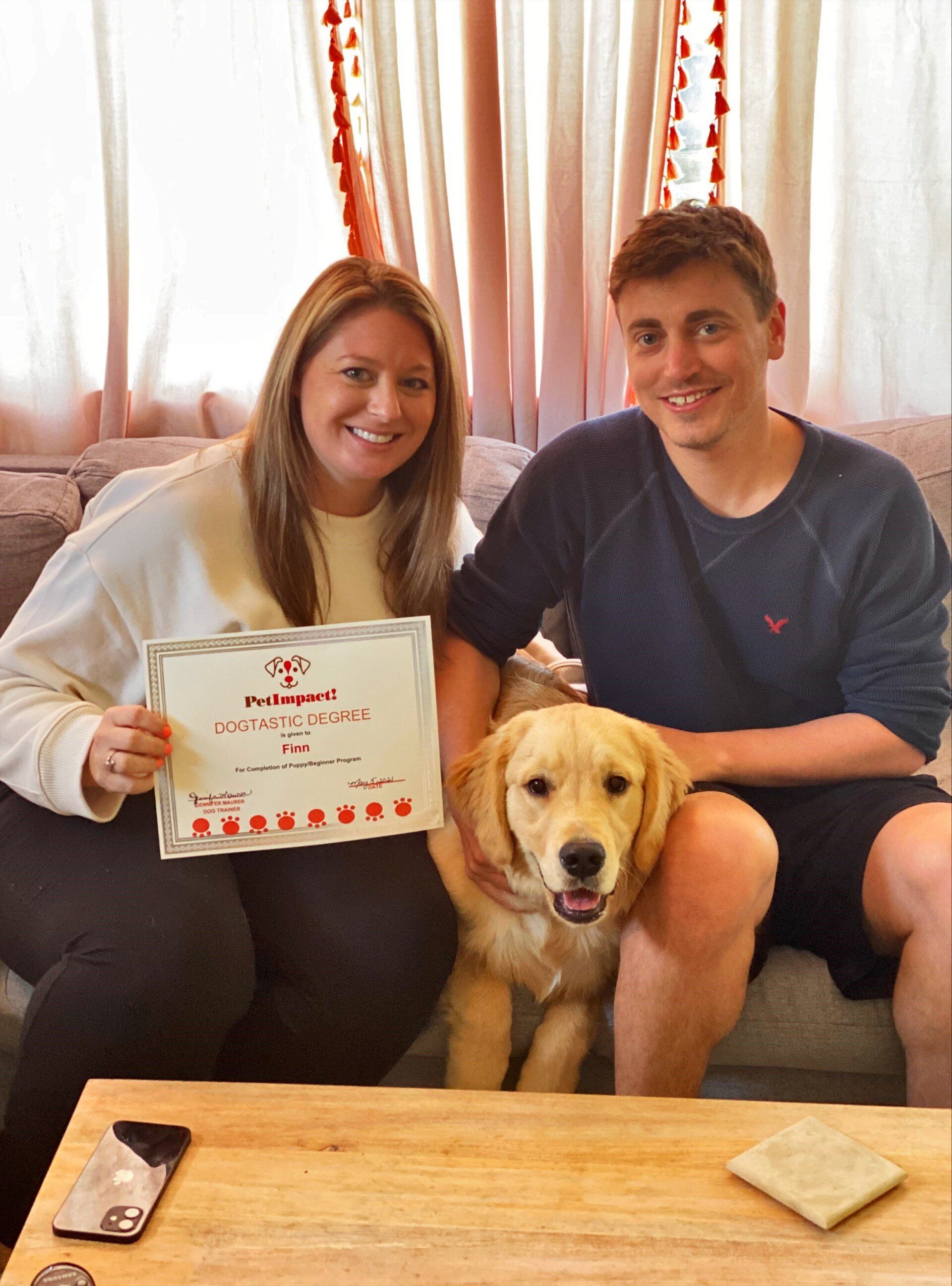 Woman and man with golden retriever puppy, holding certificate. Sitting on couch; neutral tones.