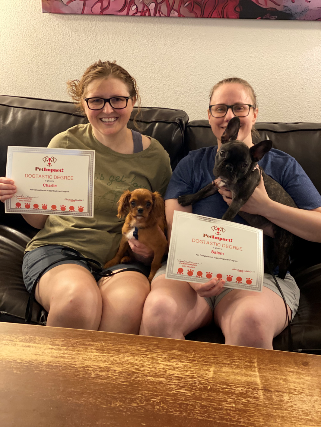 Two women on a couch with dogs, each holding a certificate. They are smiling.