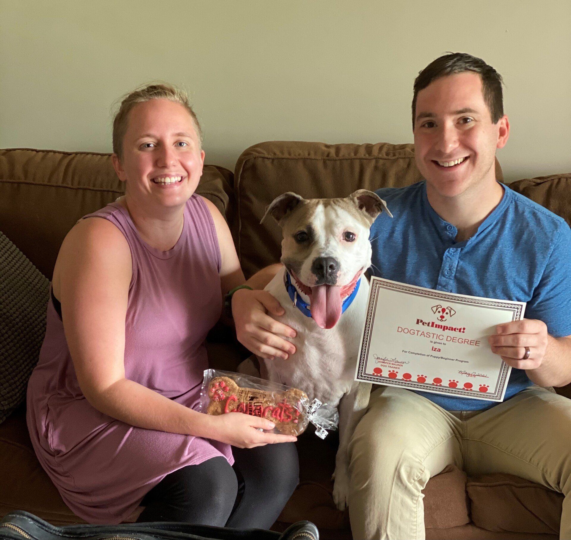 Couple and dog on couch, holding treats and certificate. Woman in pink tank, man in blue, dog smiles.