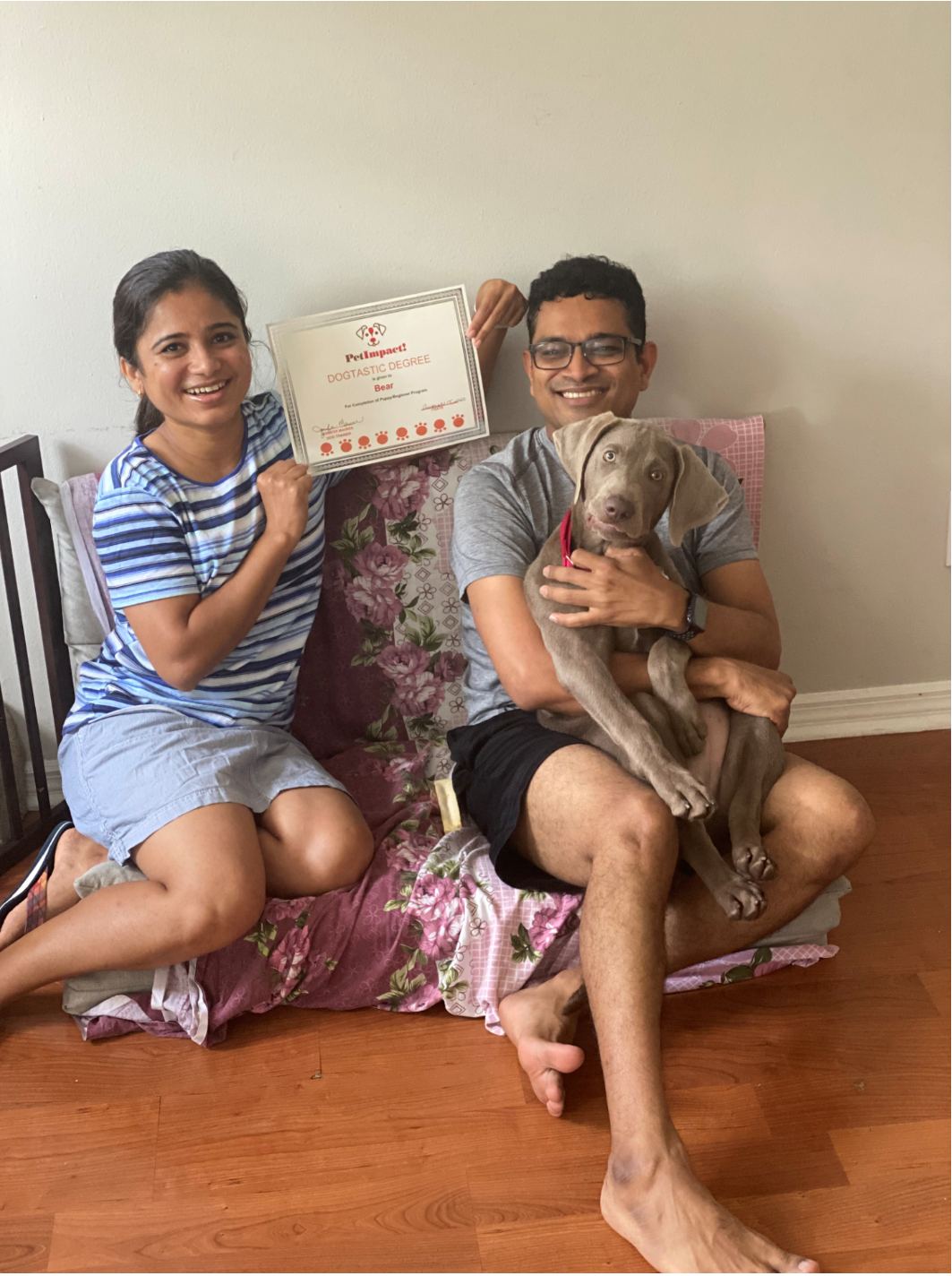 Couple with puppy and certificate, sitting on floral cushion indoors. Woman clenches fist, man holds puppy.