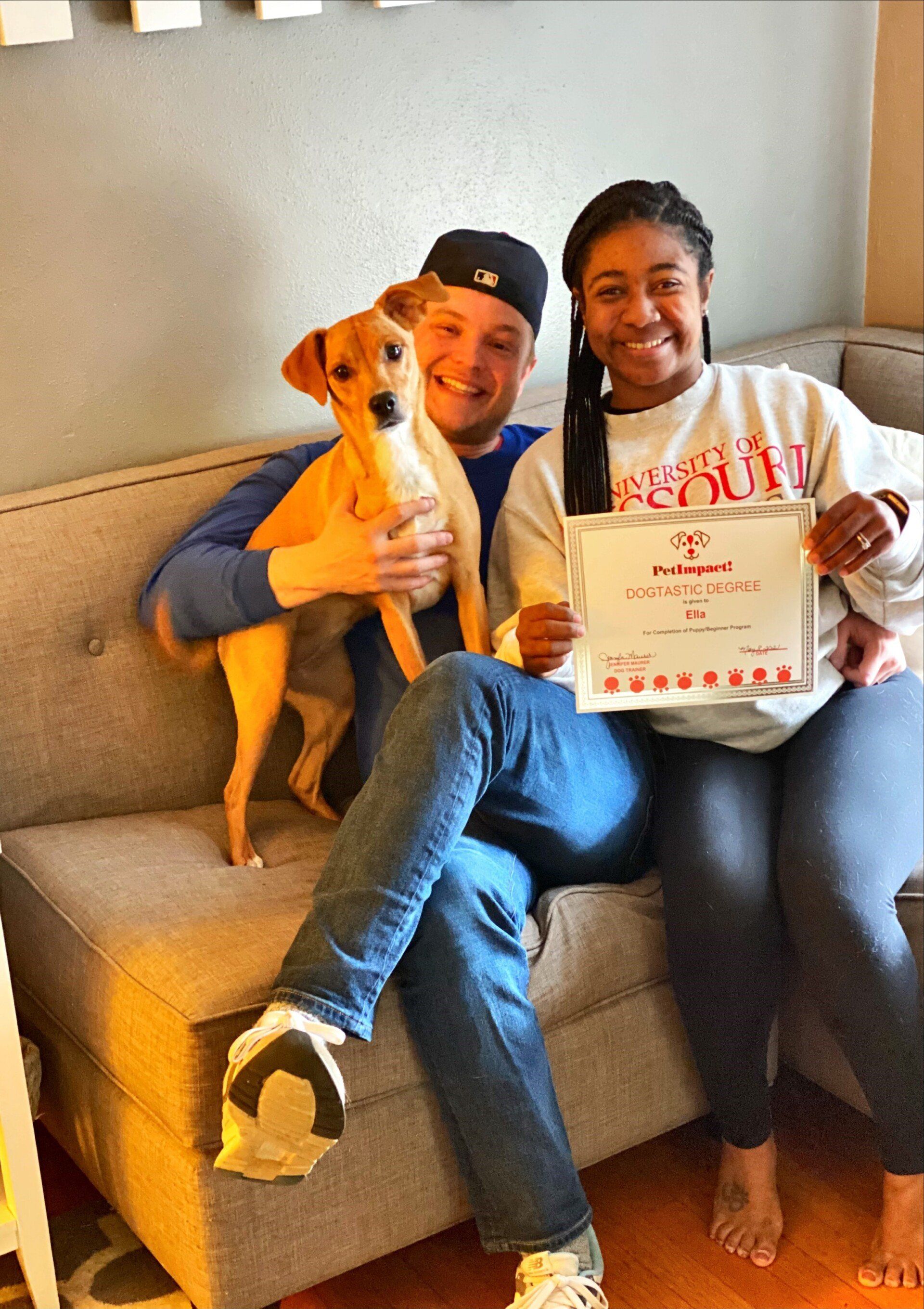 A couple sits on a couch, holding a dog and a framed certificate, smiling indoors.