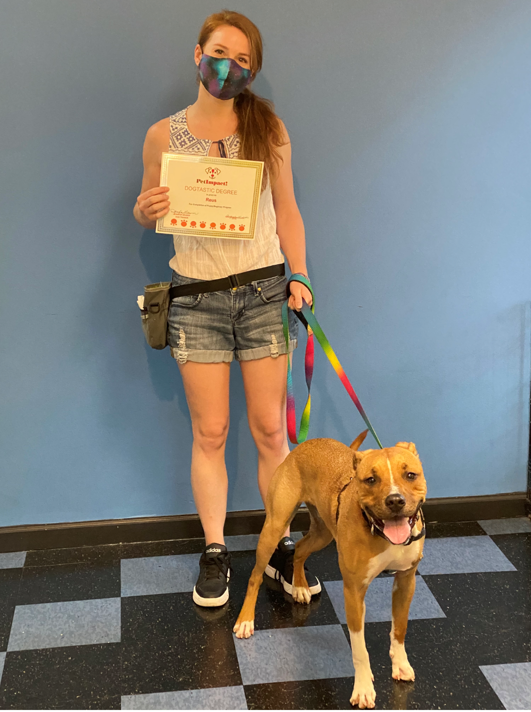 Woman in shorts holding certificate, smiling dog on leash. Blue wall background.