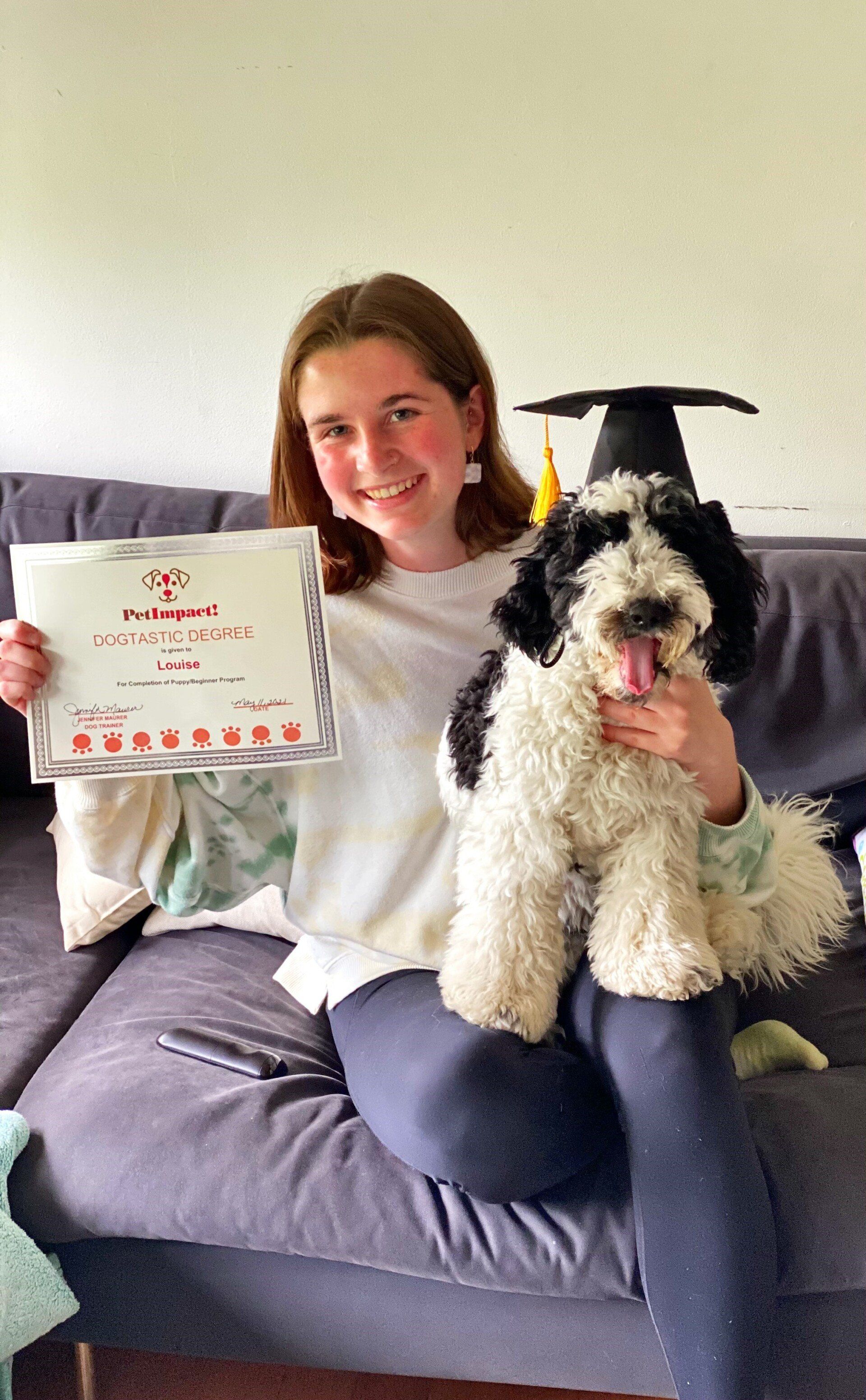 Woman smiling, holding a diploma and a fluffy black and white dog. Black graduation cap on the couch.