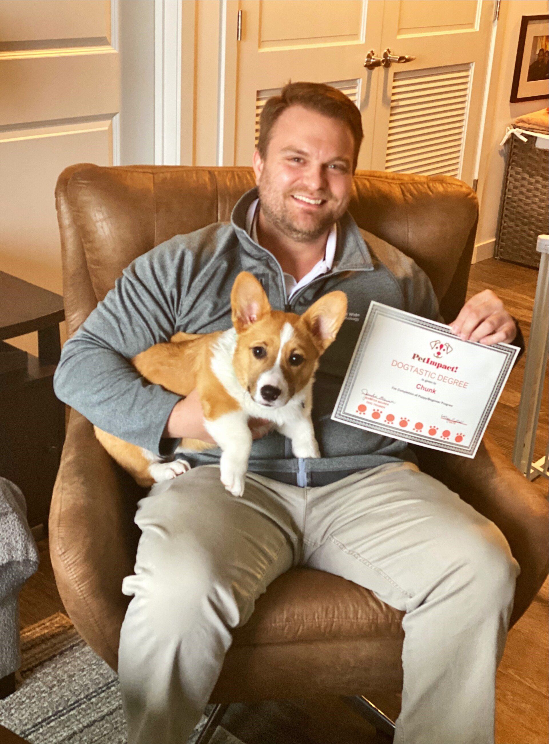 Man sitting in a chair holding a corgi and a certificate. Smiling. Indoors.