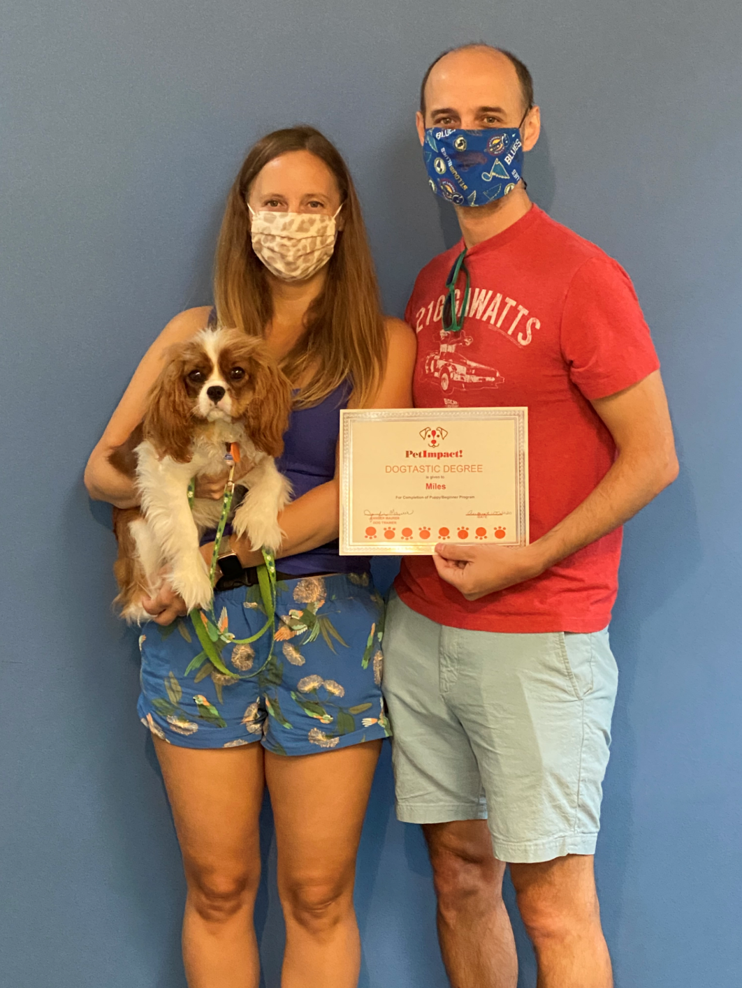 Couple and dog holding certificate; woman in blue top and shorts holds Cavalier King Charles Spaniel. Man in red shirt holds certificate.