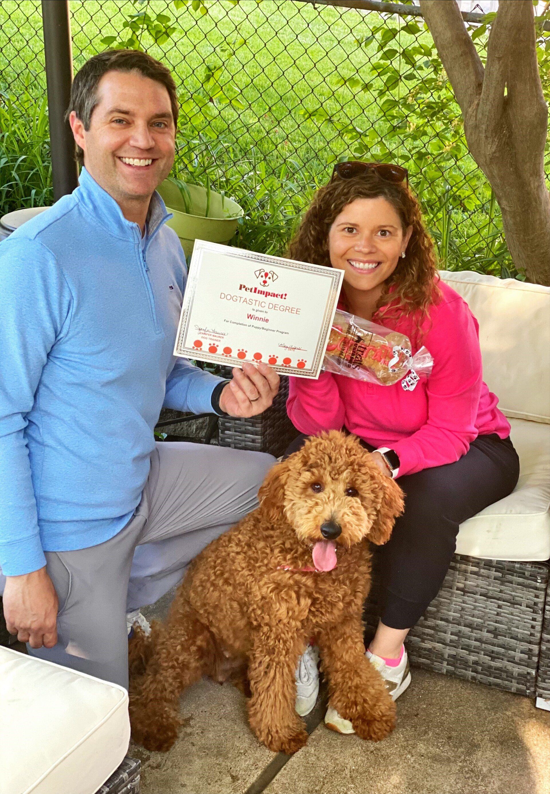 Man and woman with a dog holding a certificate and flowers, smiling outdoors.