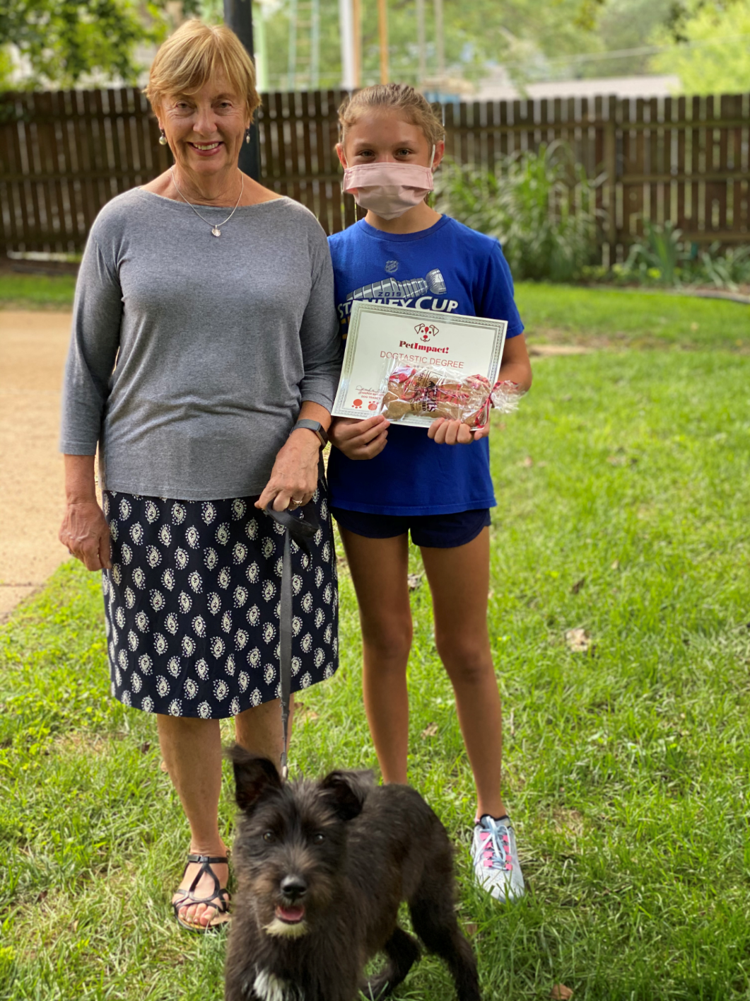 Woman and girl with a dog, standing outside. Girl holds a printed paper, wearing a mask.
