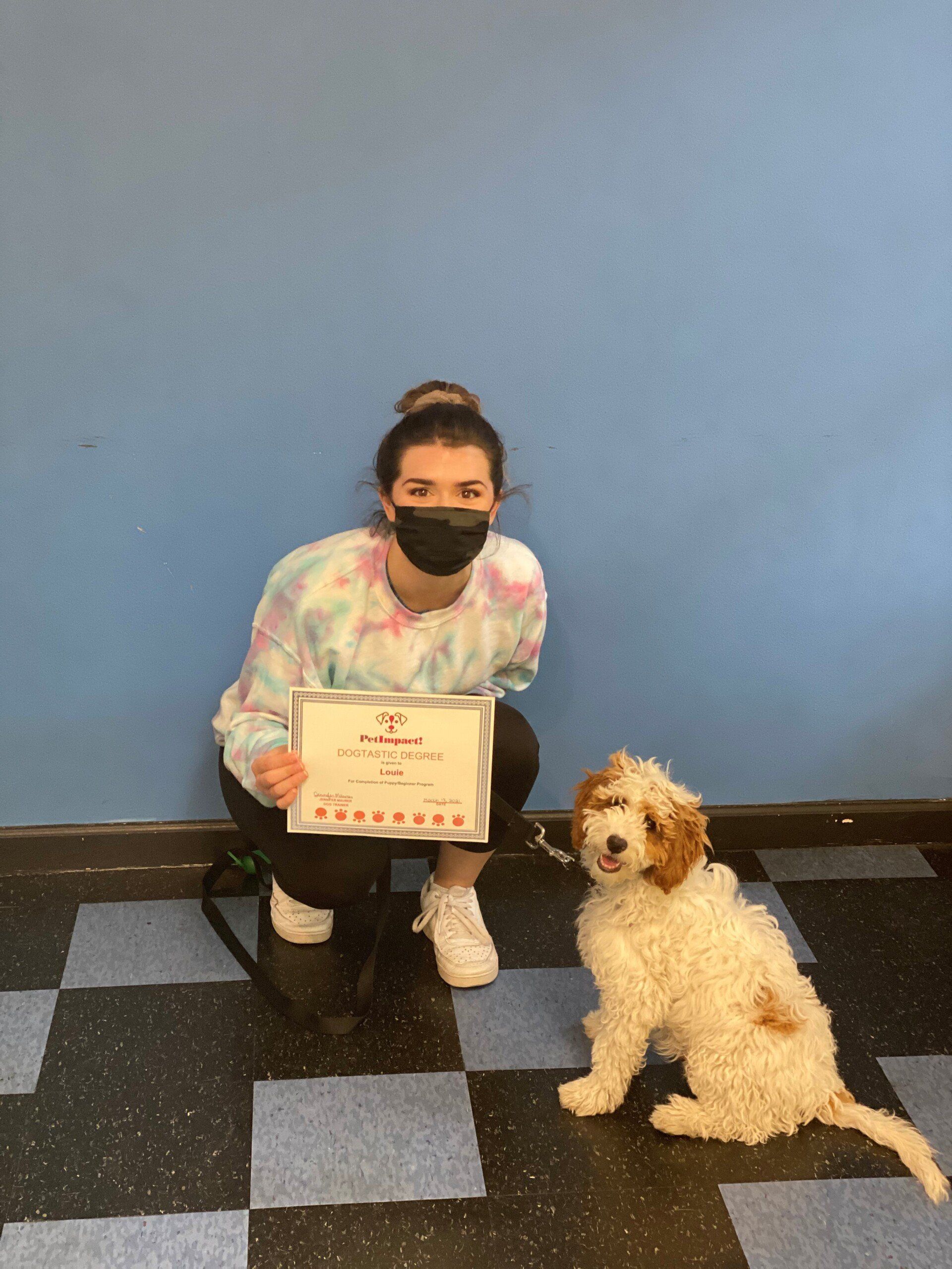 Woman wearing a mask and tie-dye shirt kneels with a curly-haired dog holding a certificate in front of a blue wall and checkered floor.