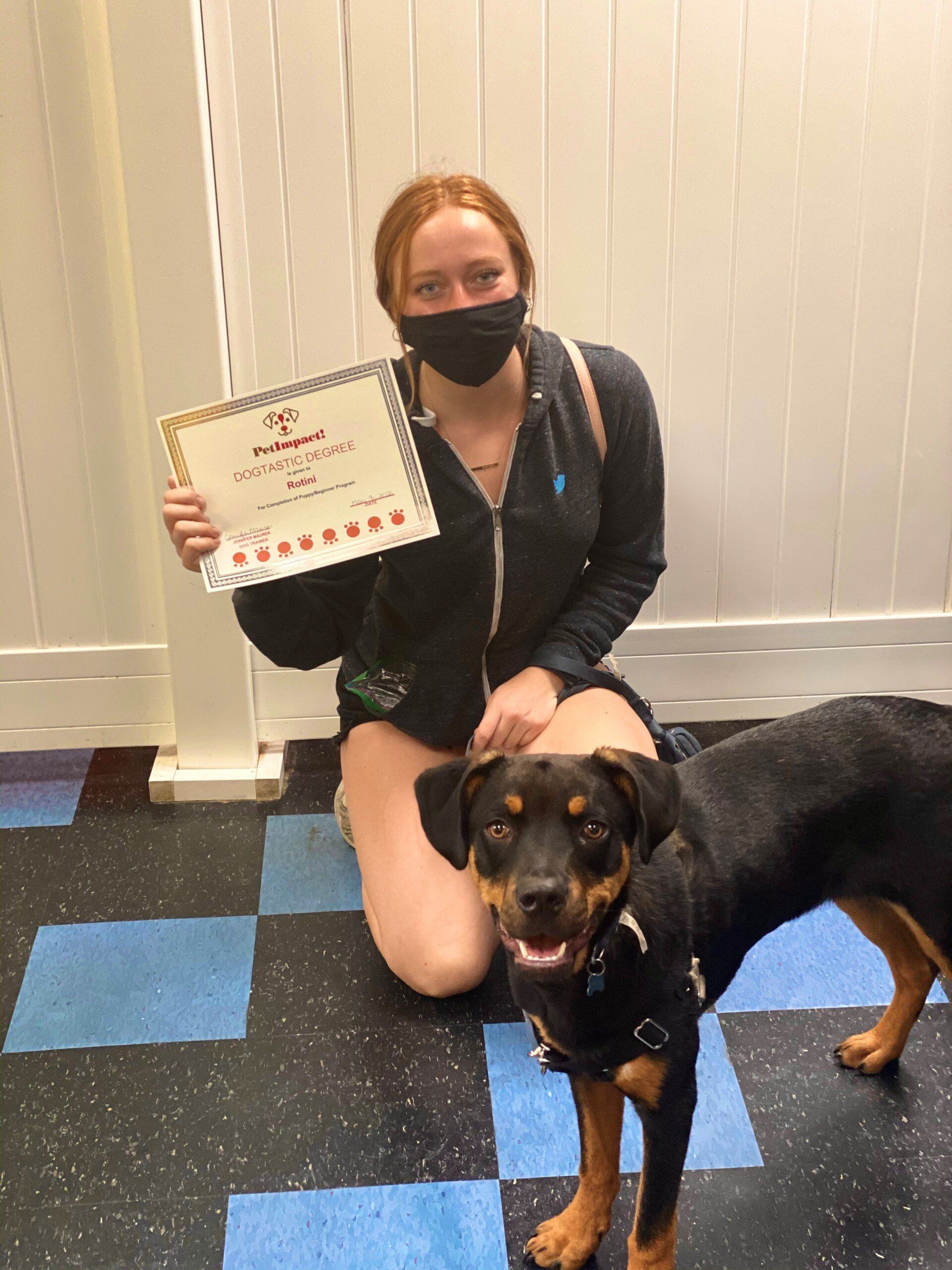 Woman in black mask kneels with dog, holding certificate.  They are in a room with a blue and black tiled floor.