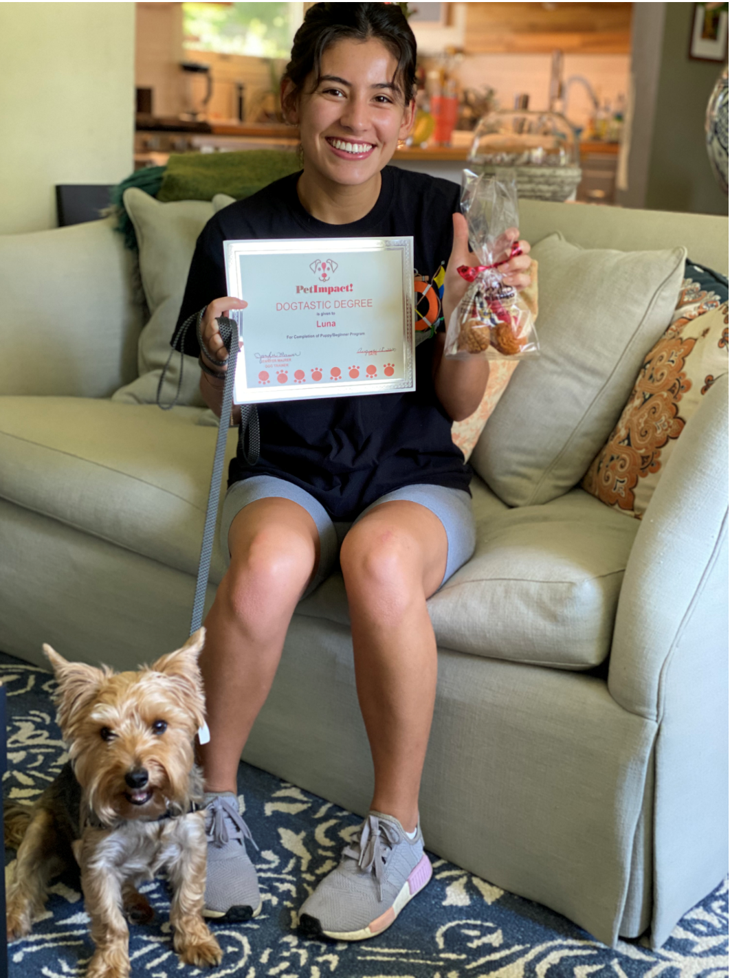 Woman on couch holding a certificate and treats, with a small dog on a leash.