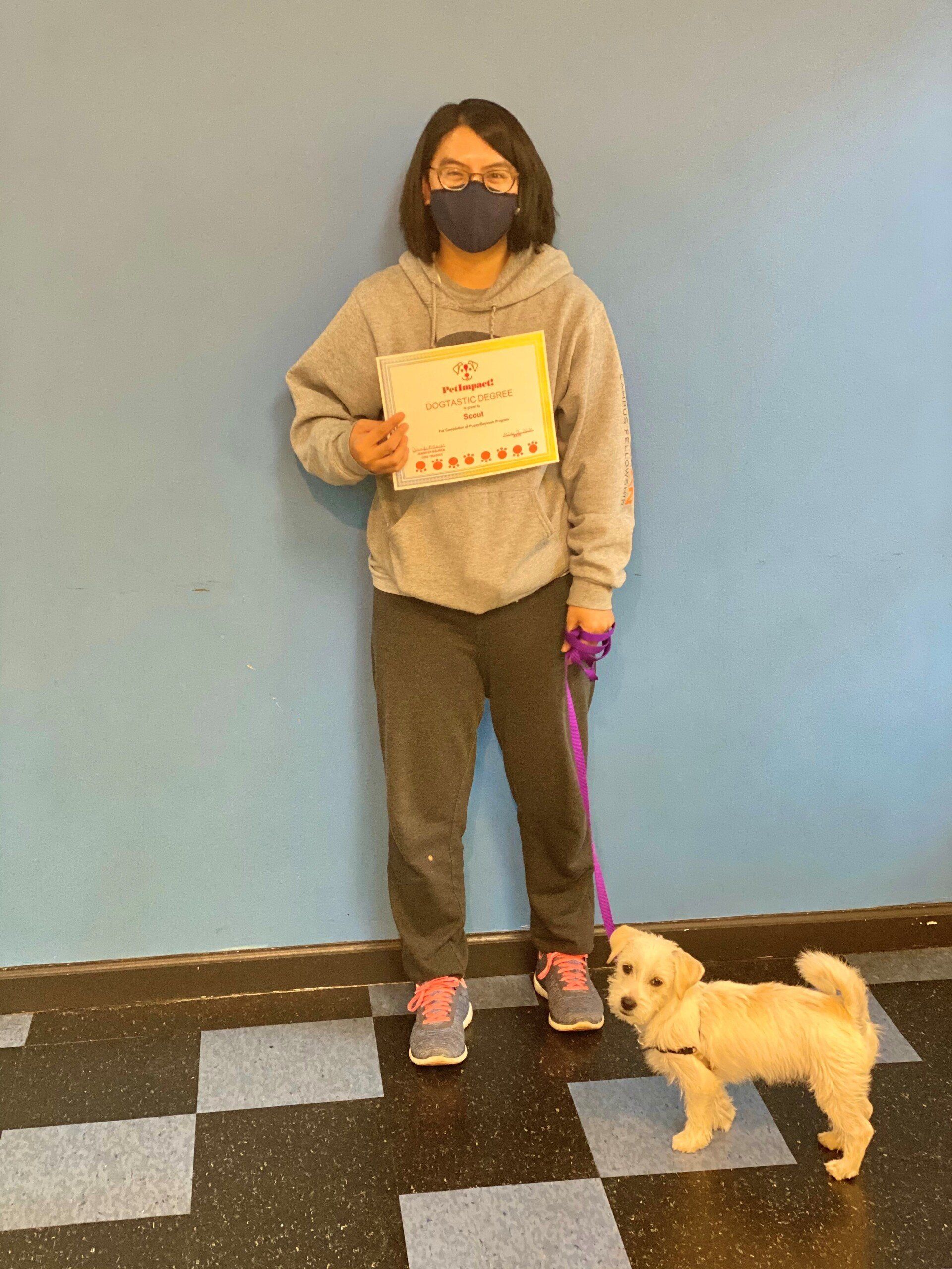 Person in gray sweatsuit and mask holding a certificate, standing with a small white dog on a leash in front of a blue wall.
