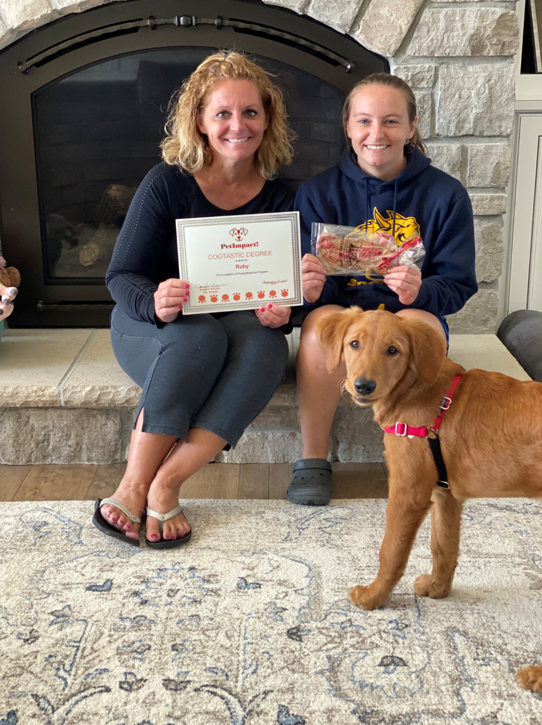 Woman and girl with a golden retriever pose near fireplace. They hold certificate and treat bag.