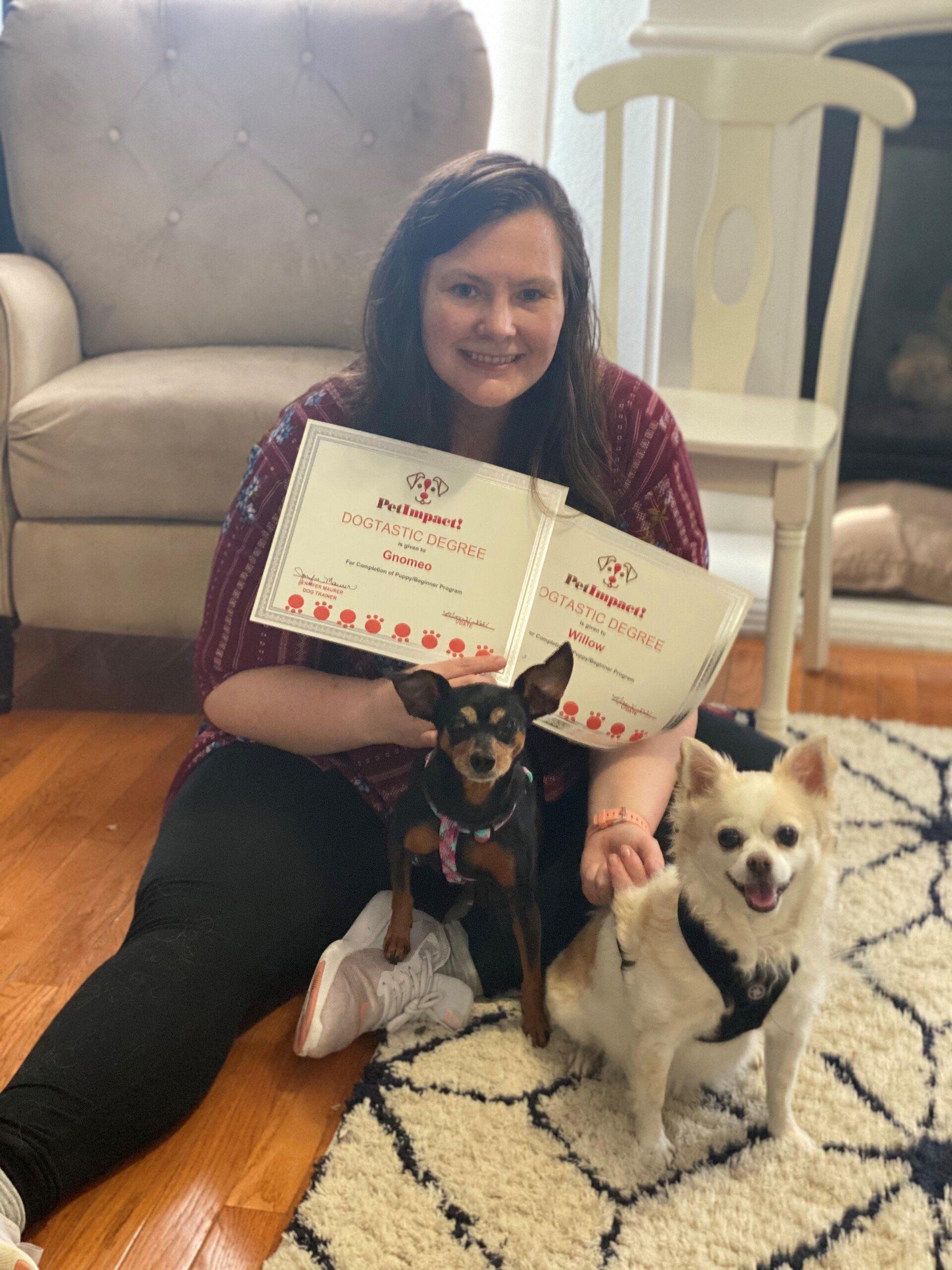 Woman sits with two dogs holding certificates on a rug. Smiling, brown hair.