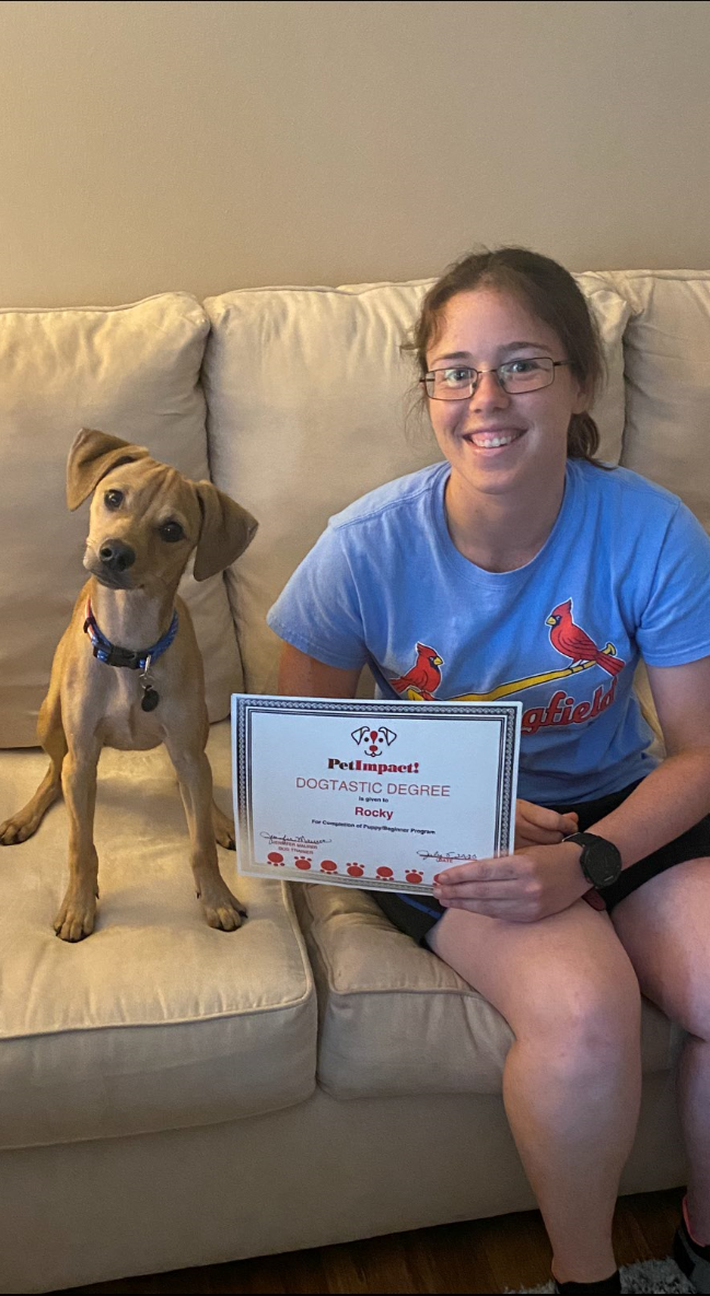 Woman and dog on a couch holding a certificate, both smiling. Dog has a tilted head.