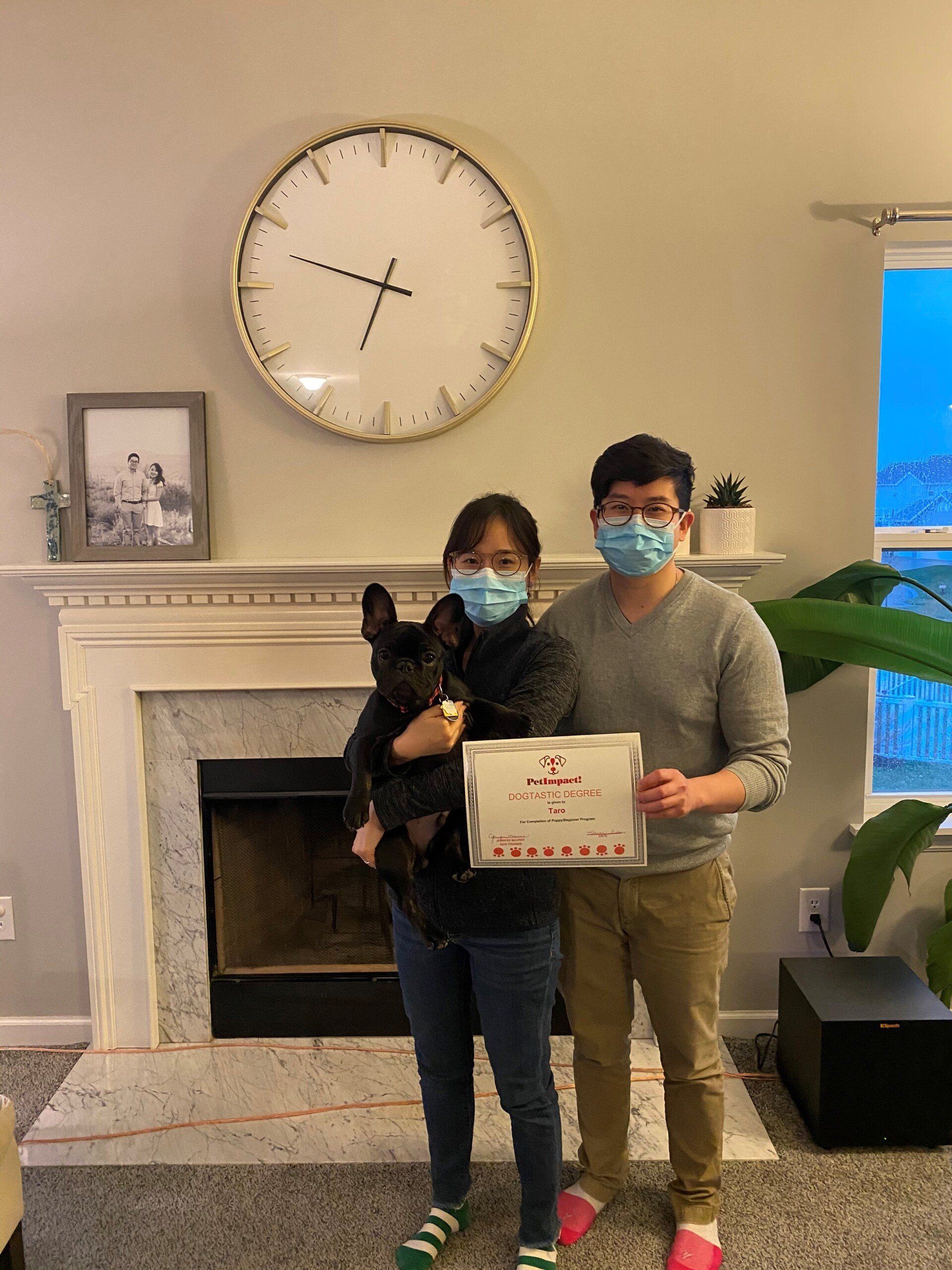 Couple, dog, and certificate in front of a fireplace. They all wear masks. A large clock and photo hang on the wall.