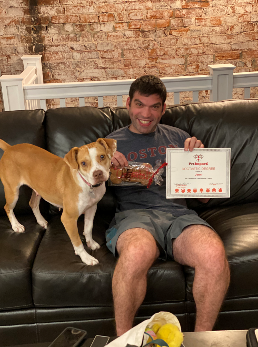 Man on black couch with dog, holding baked goods and a certificate, smiling. Brick wall background.