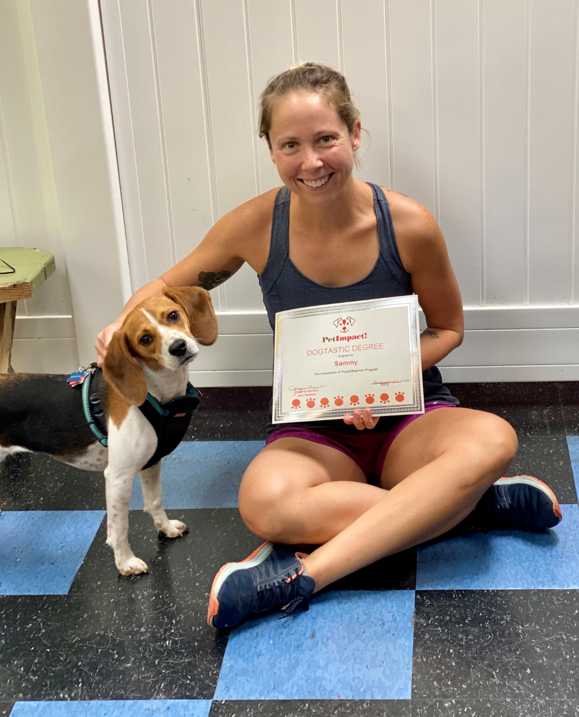 Woman and beagle sit together, holding award. Woman smiles, dog looks at camera. They're on a checkered floor.