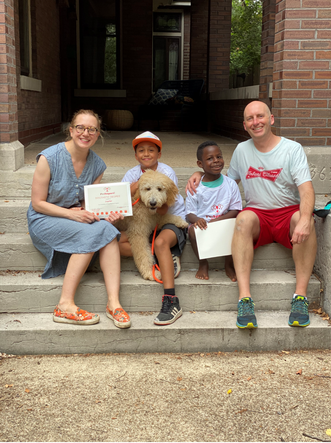 Family and dog pose on steps; woman in blue dress holds certificate; children, man in red shorts smile.
