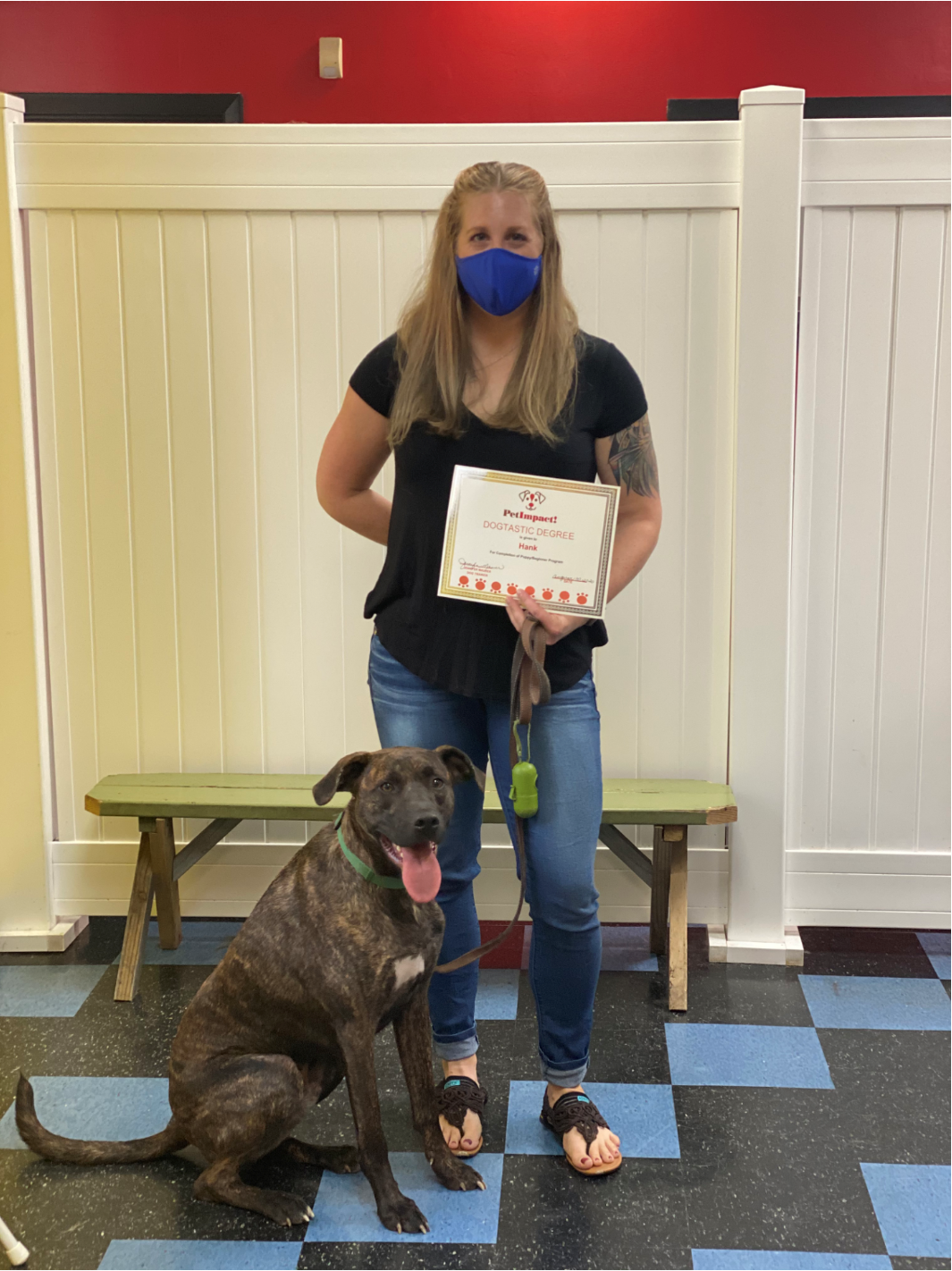 Woman in mask holds certificate with dog sitting by her feet, inside a facility.