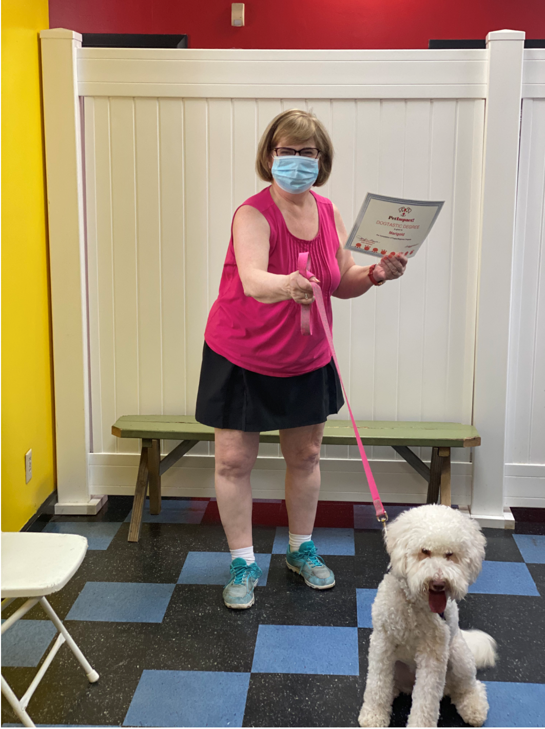 Woman in pink top and mask holds a certificate and leash, standing with a white dog in a room.