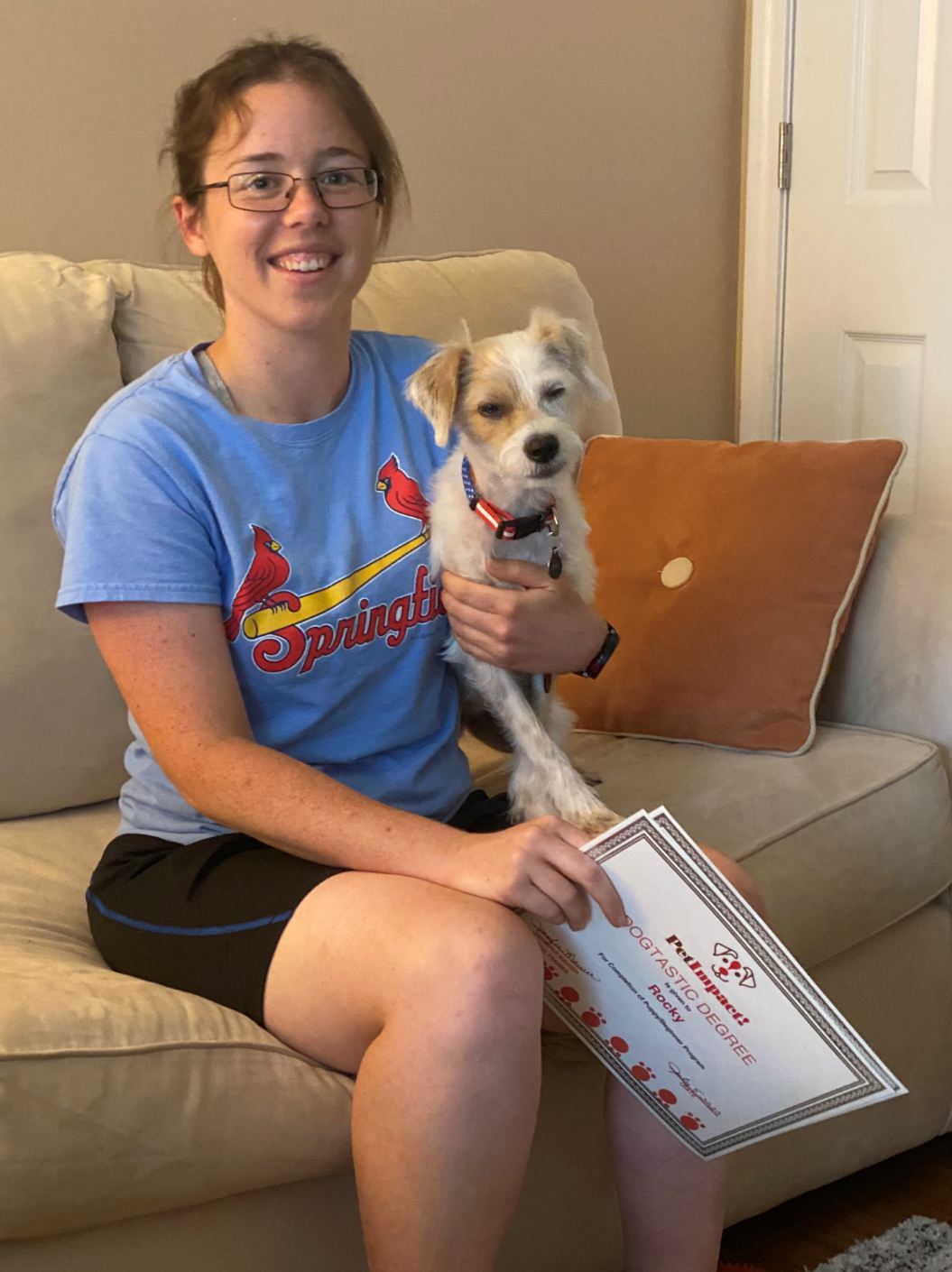 Woman on couch smiles, holding small dog and a certificate.