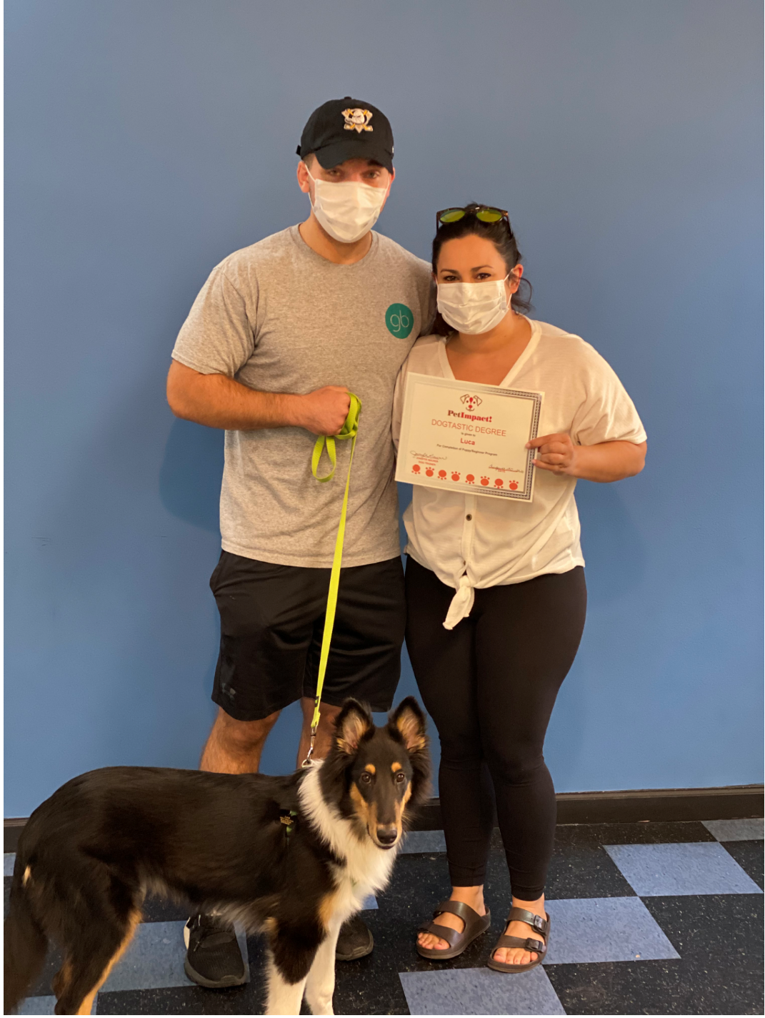 Man and woman with dog, both wearing masks, hold a certificate in front of a blue wall.