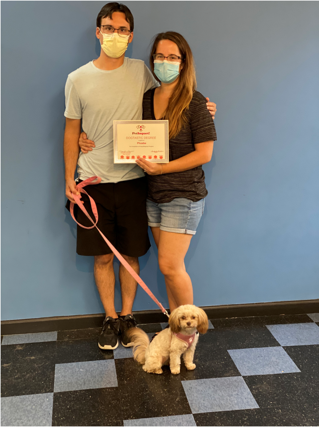Couple with small dog in front of blue wall. They hold a certificate and wear masks. Dog on leash.