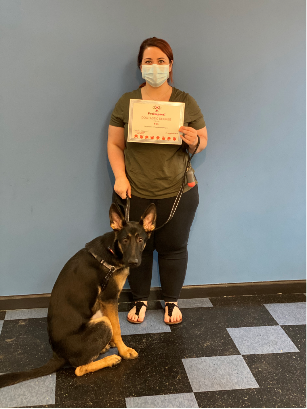 Woman in face mask with dog, holding certificate in front of a blue wall and checkered floor.