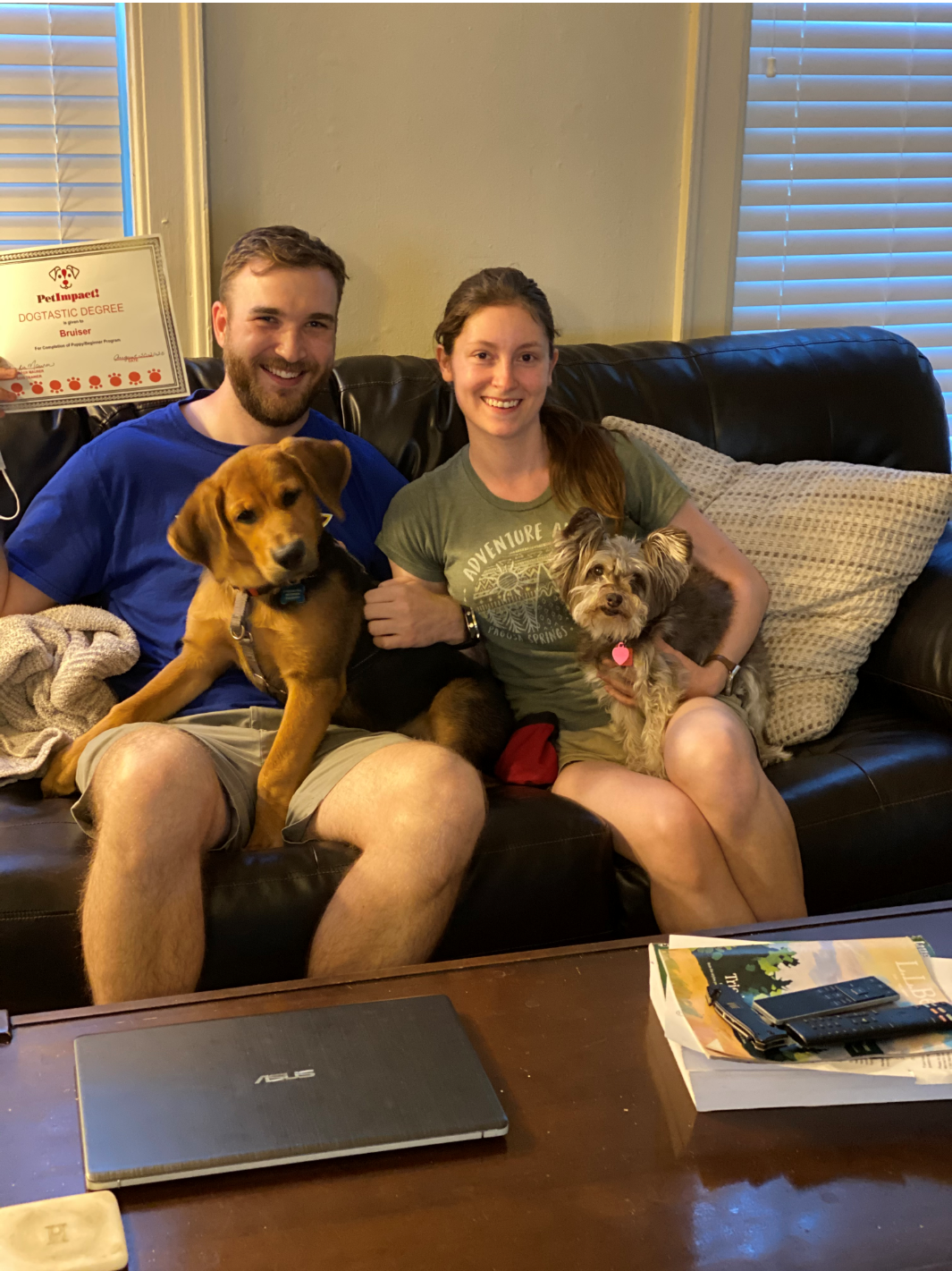 Couple on a sofa with two dogs; man holds a brown dog, woman holds a gray dog. Indoors.