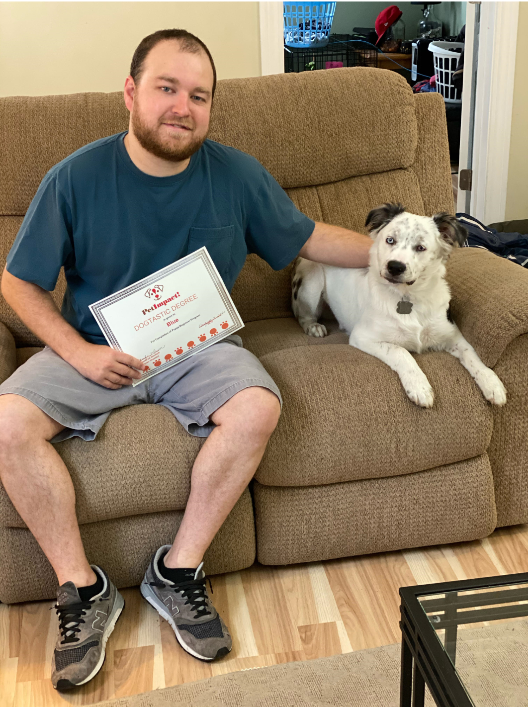 Man and dog on a couch. Man holds certificate, smiles, and pets the dog. The dog is white with black spots.
