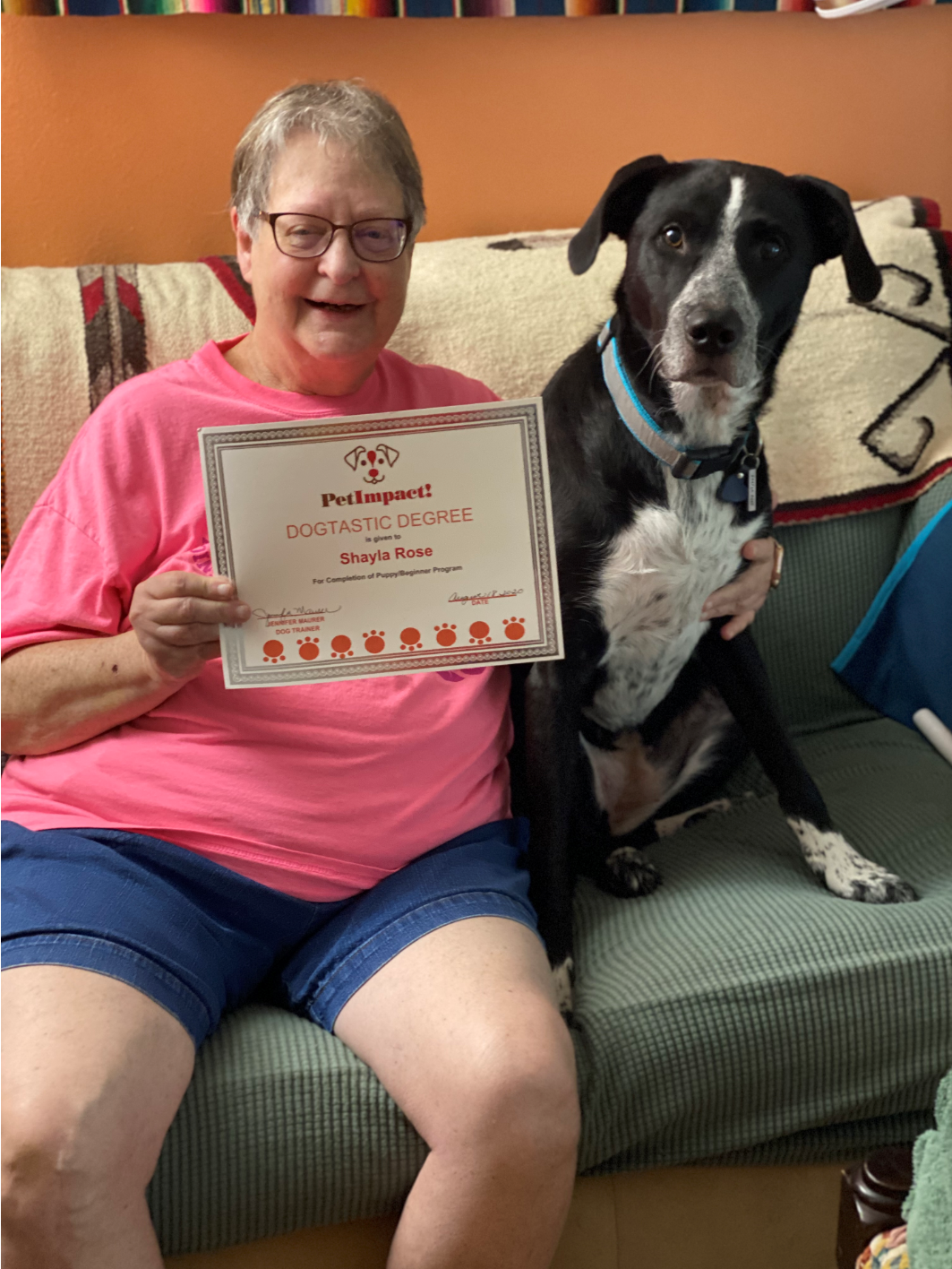 Woman and dog on a couch, holding a certificate. The dog is black and white, and the woman is wearing pink and blue.