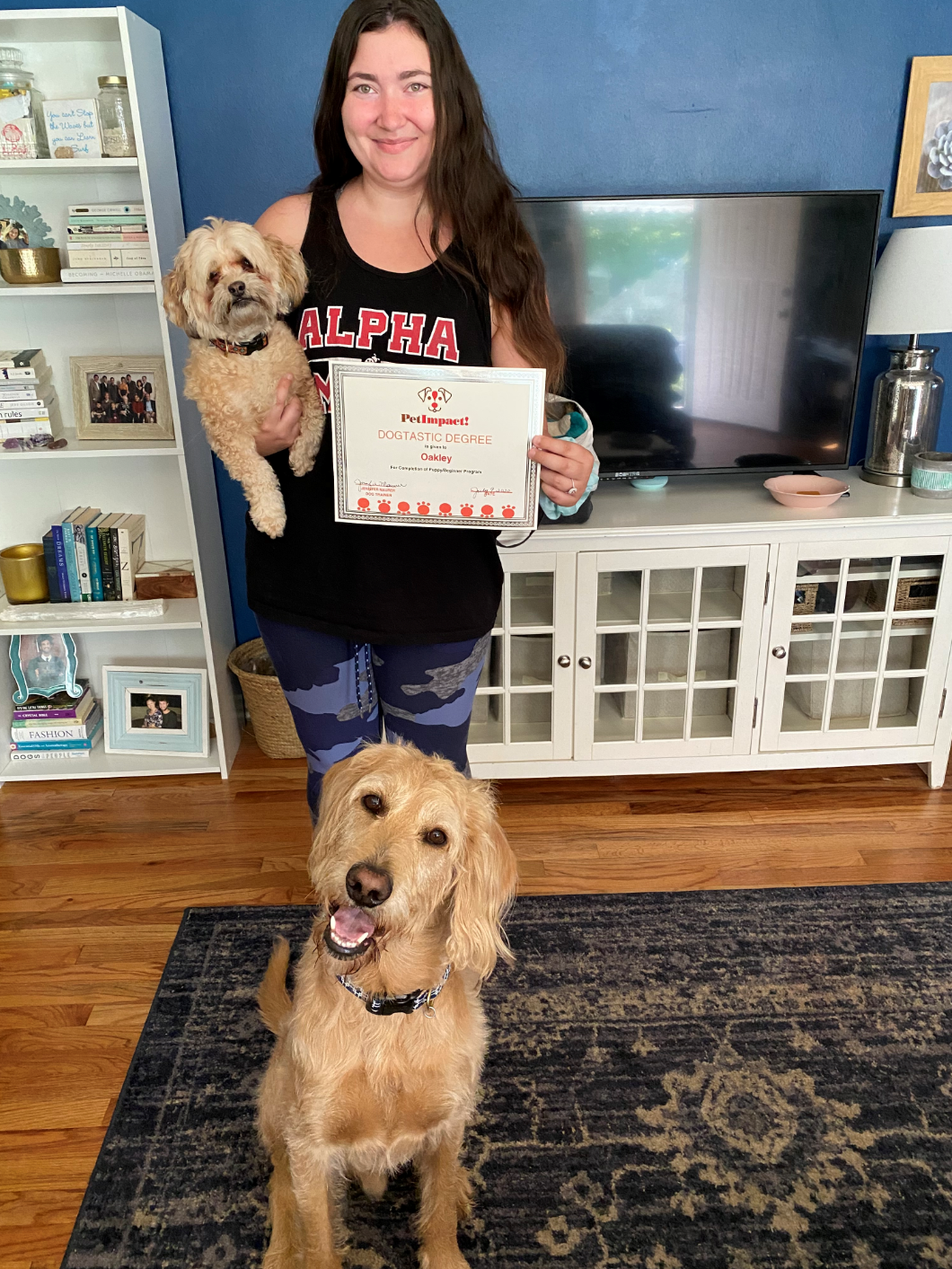 Woman holding certificate and small dog, larger dog sits on rug. Home interior, smiles.