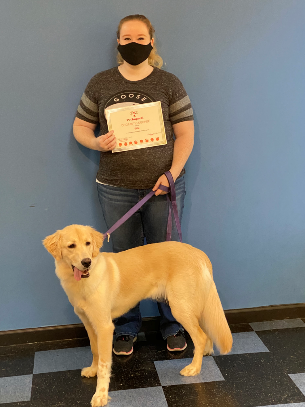 Woman wearing mask holds certificate, stands with a golden retriever on a leash in front of a blue wall.