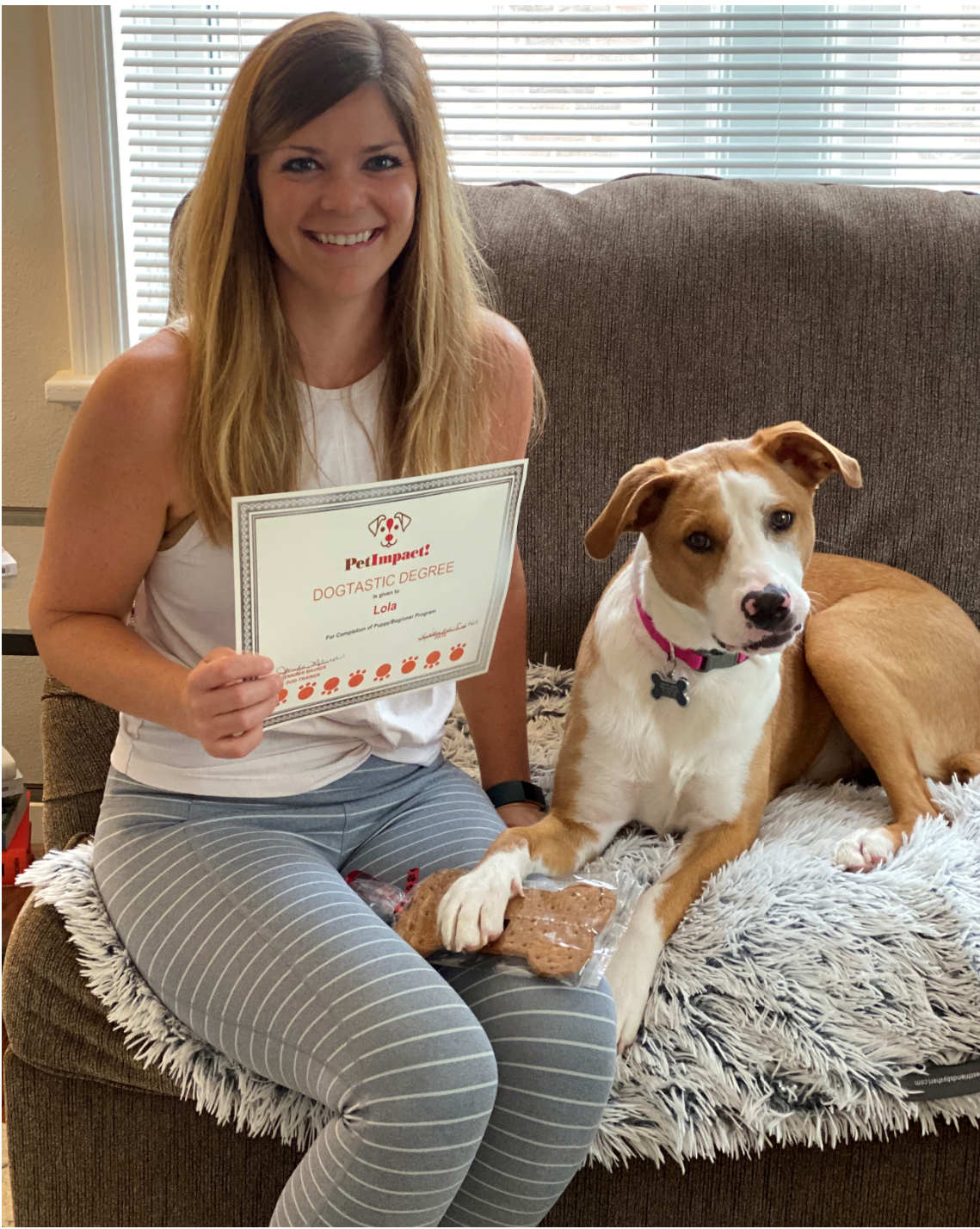 Woman and dog on a couch holding a certificate and dog treat. Both are smiling.